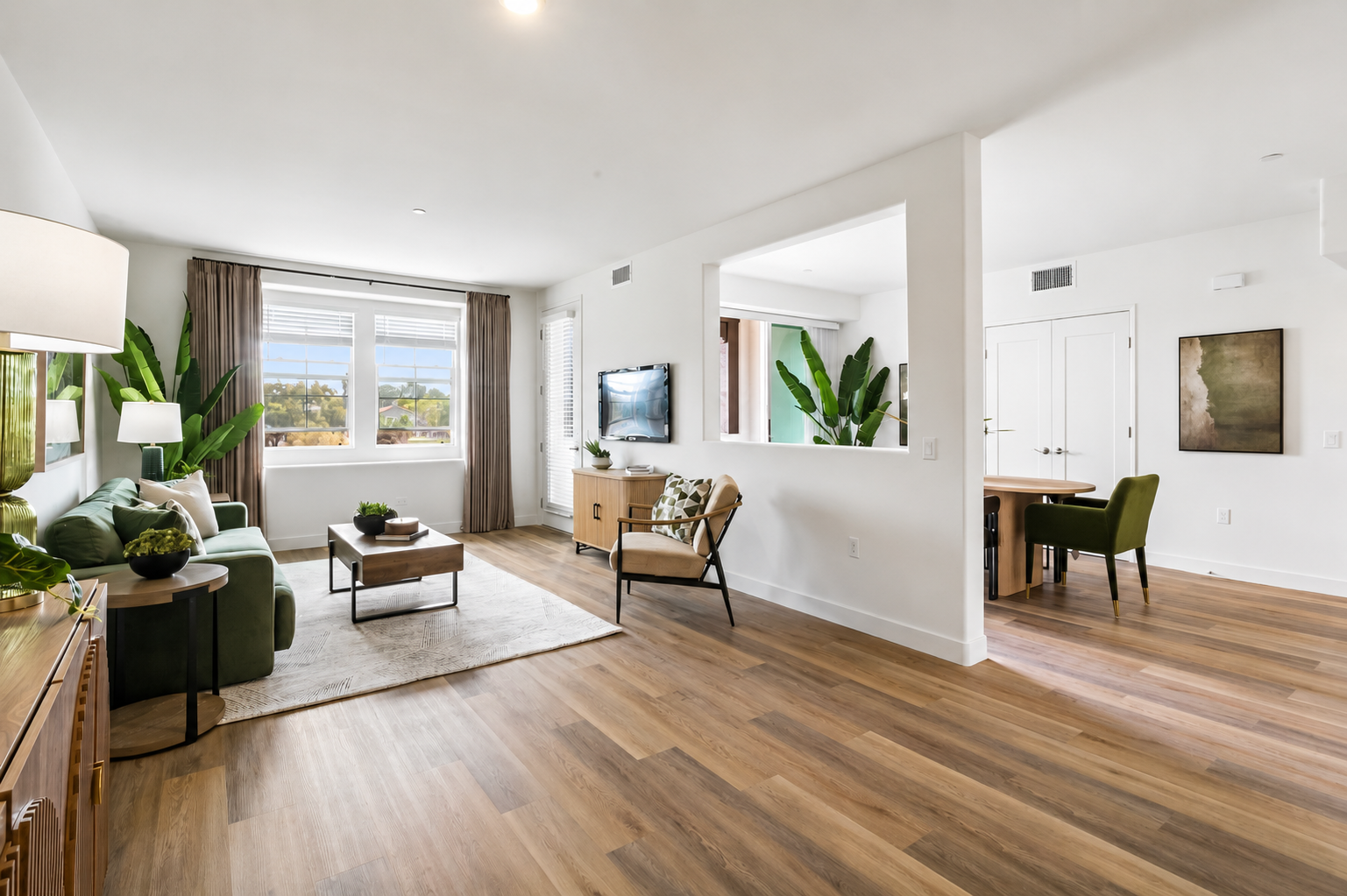 A modern, stylish living room featuring a green sofa, a wooden coffee table on a light rug, and large windows with natural light. Potted plants add greenery to the space. The room transitions into a dining area with a small table and chairs, and wooden flooring enhances the cozy atmosphere.