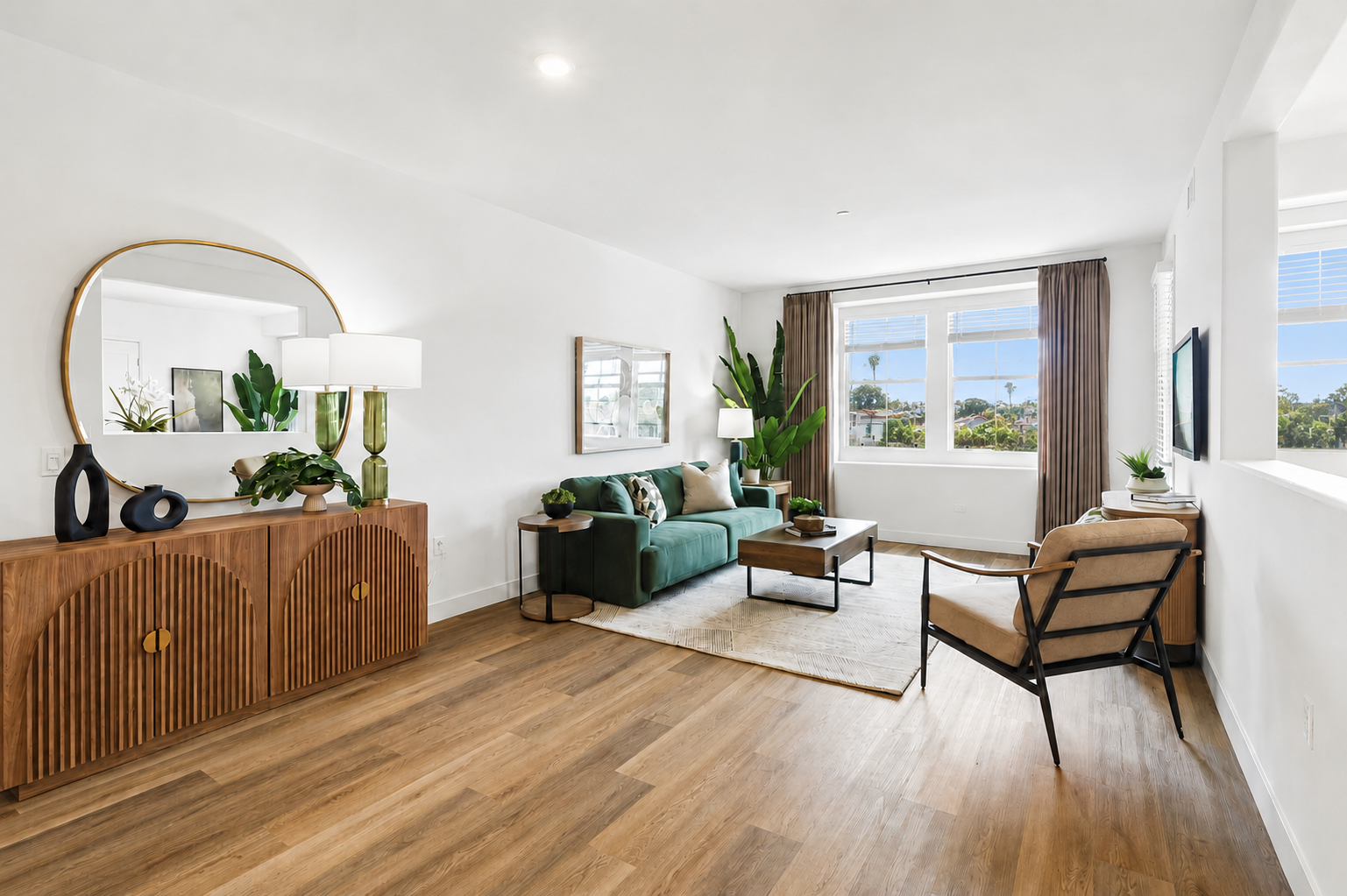 A modern living room featuring a green sofa, decorative pillows, a wooden side table, and a large round mirror. Natural light enters through a window with sheer curtains, illuminating the space. Indoor plants add a touch of greenery, complemented by a neutral rug on a wooden floor.