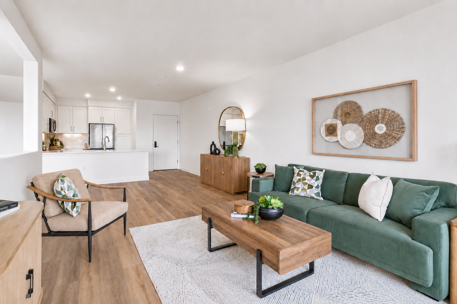 A modern living room featuring a green sofa with decorative pillows, a wooden coffee table, and a light-colored area rug. There are two chairs, a side table, and a large round mirror on the wall. The kitchen area is visible in the background with white cabinetry and appliances. Decorative art pieces are displayed on the walls.