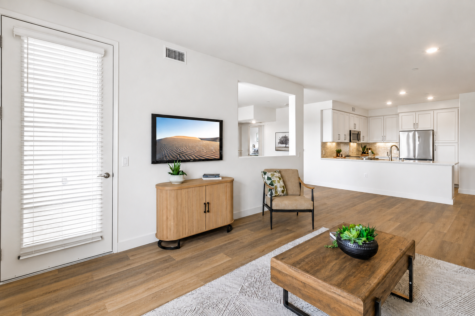 A modern living room featuring light-colored walls, wooden flooring, and minimalist decor. There's a TV mounted on the wall displaying a desert scene, a wooden coffee table with a plant, and an armchair. The space is open, leading to a kitchen area with sleek cabinets and appliances in the background. Natural light comes through a window with shutters.