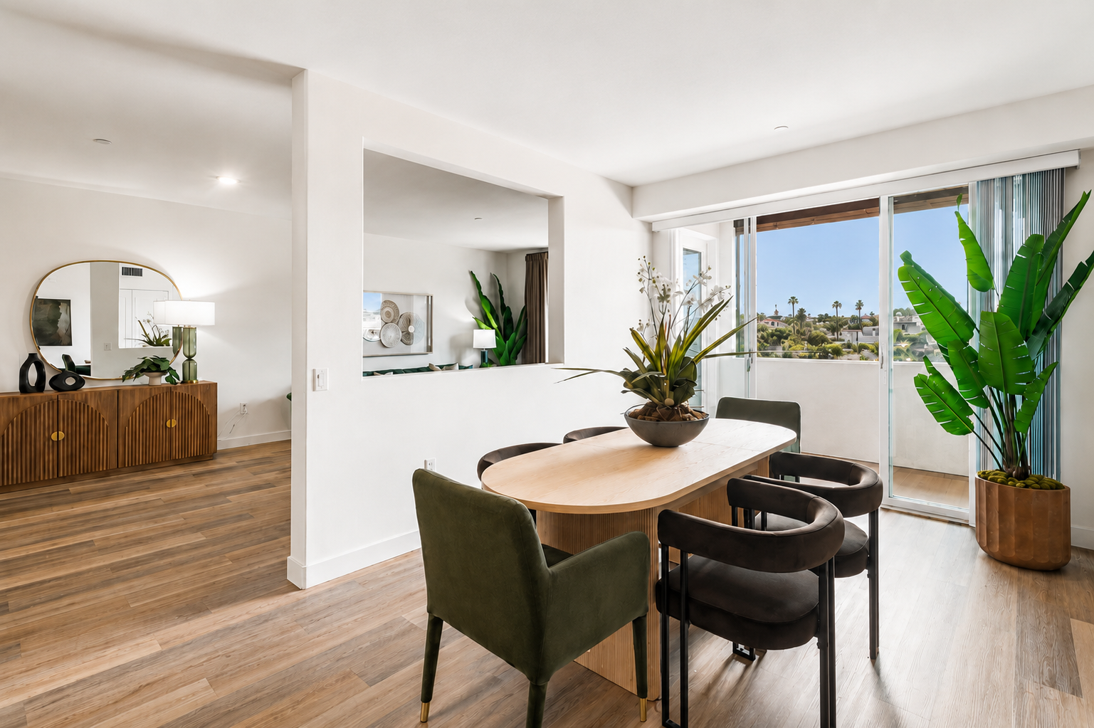 A modern dining area featuring a round wooden table surrounded by green and black chairs. In the background, there is a stylish console table with decor, and large windows with a view of greenery outside. Bright, natural light floods the space, highlighted by indoor plants and contemporary furnishings.