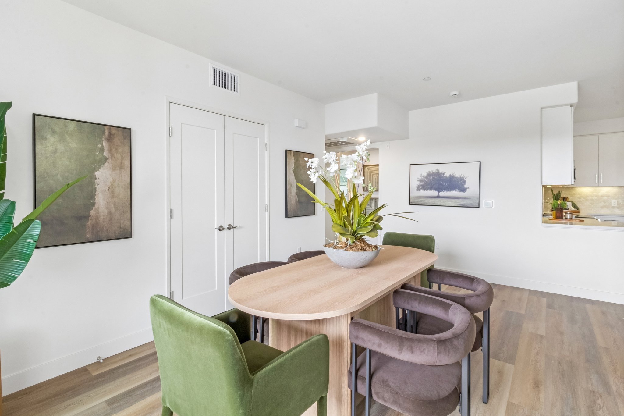 A modern dining area featuring a round wooden table surrounded by green and brown upholstered chairs. Wall art in neutral colors adorns the walls, and a potted orchid centerpiece adds a touch of elegance. The space is bright and airy, with an open kitchen visible in the background.