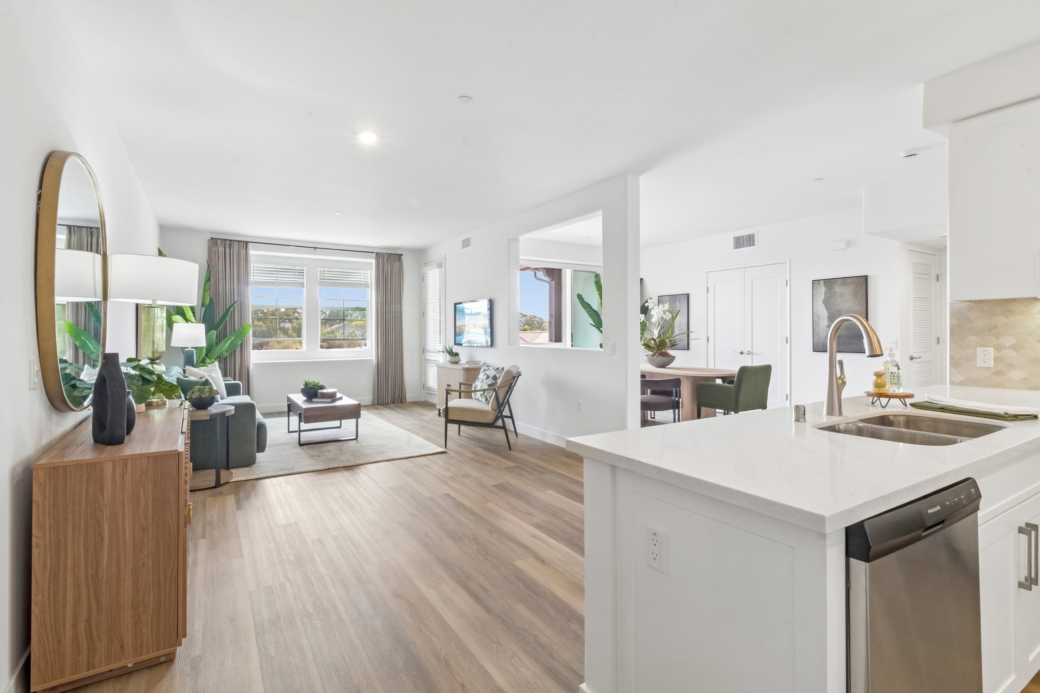 A modern, open-concept living space featuring a kitchen with a stainless steel dishwasher. The room includes a stylish dining area, comfortable seating, large windows with views, and decorative plants. Light wood flooring complements the contemporary decor, creating a bright and inviting atmosphere.