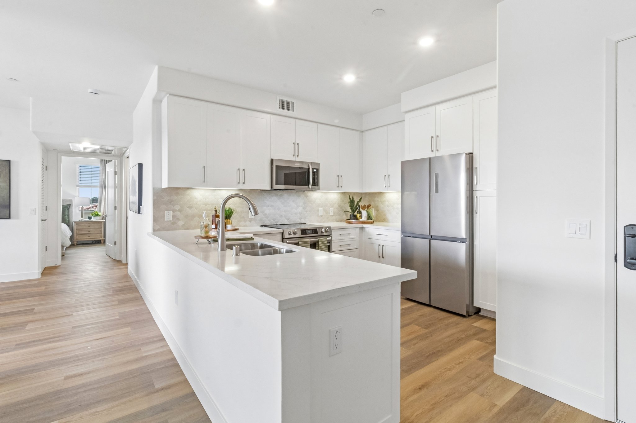 Modern kitchen with white cabinetry, stainless steel appliances, and a marble countertop. Features a spacious layout with a large sink, stove, and refrigerator. Bright lighting enhances the clean, minimalist design. A doorway leads to another room, visible at the end of the hallway.