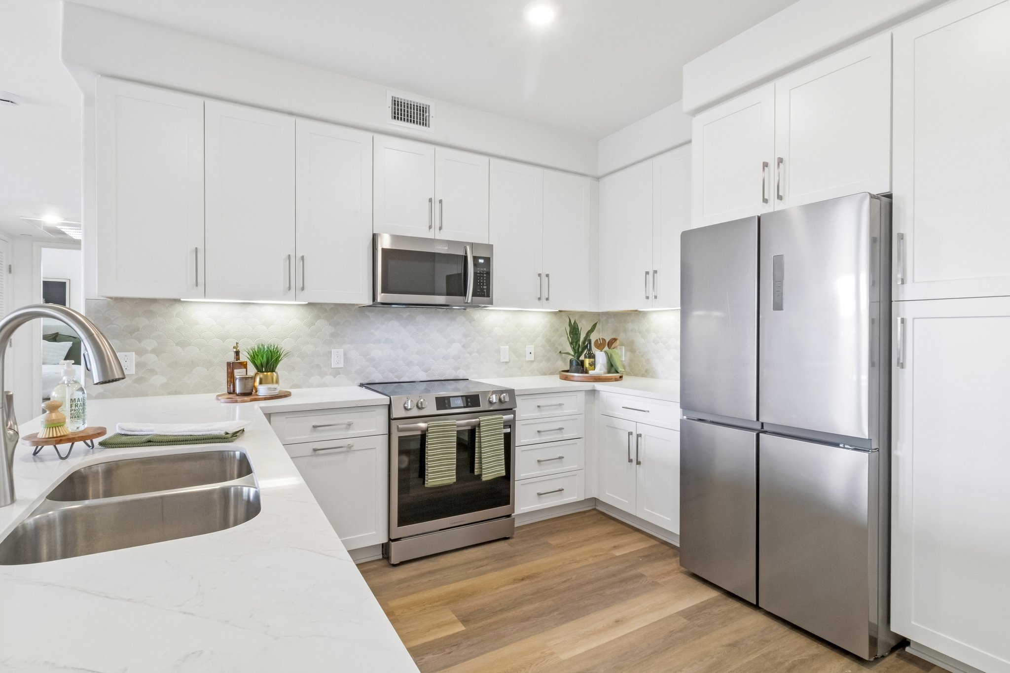Modern kitchen featuring white cabinetry, stainless steel appliances including a refrigerator and oven, a sleek countertop, and a sink with a faucet. The backsplash is composed of light-colored tiles, and there are decorative plants on the countertop, enhancing the contemporary design.