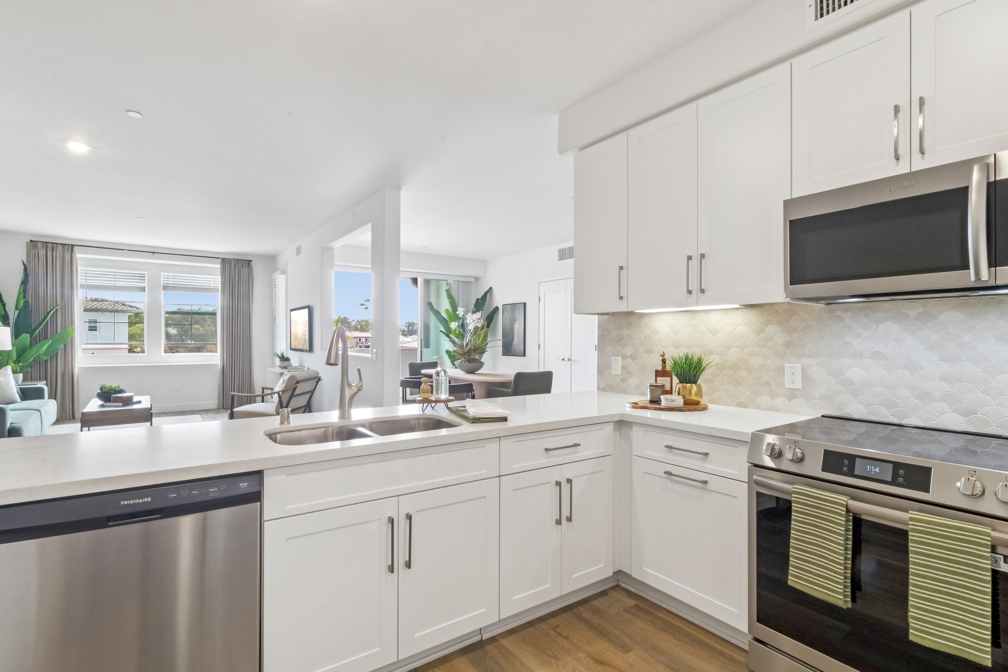 Modern kitchen featuring white cabinetry, stainless steel appliances including a dishwasher and oven, and a sleek countertop. The space is bright with natural light, showcasing a cozy living area in the background with plant accents and large windows.