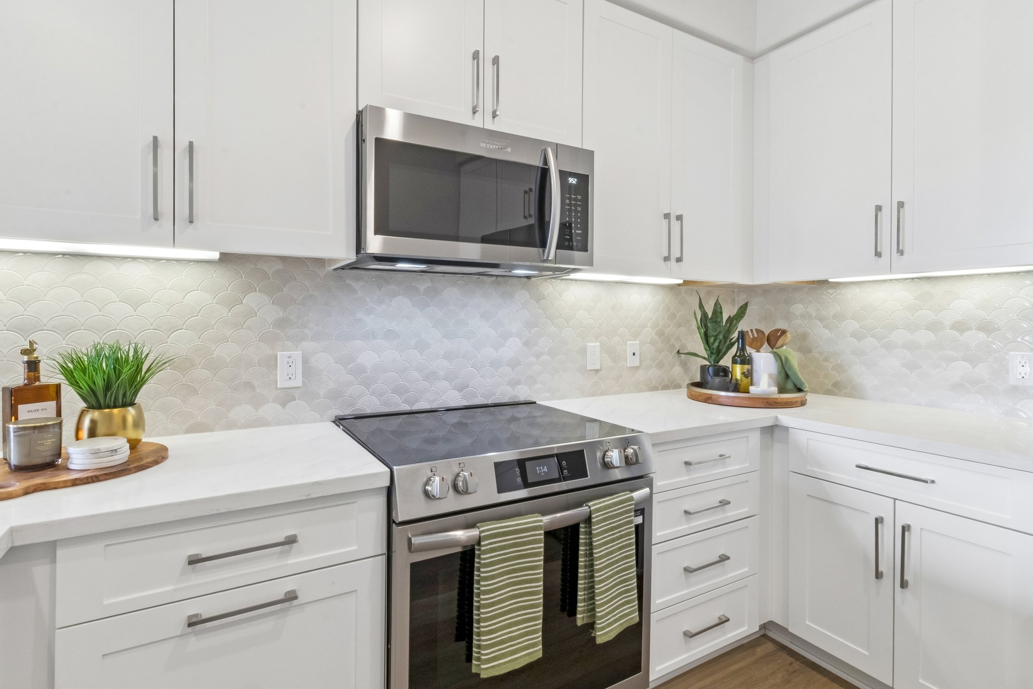 Modern kitchen featuring white cabinetry, a stainless steel microwave, and a sleek stove. Subtle gray and white scalloped backsplash complements the countertop. Wooden serving board holds kitchen utensils and plants, adding a touch of greenery and warmth to the space. Soft under-cabinet lighting enhances the ambiance.