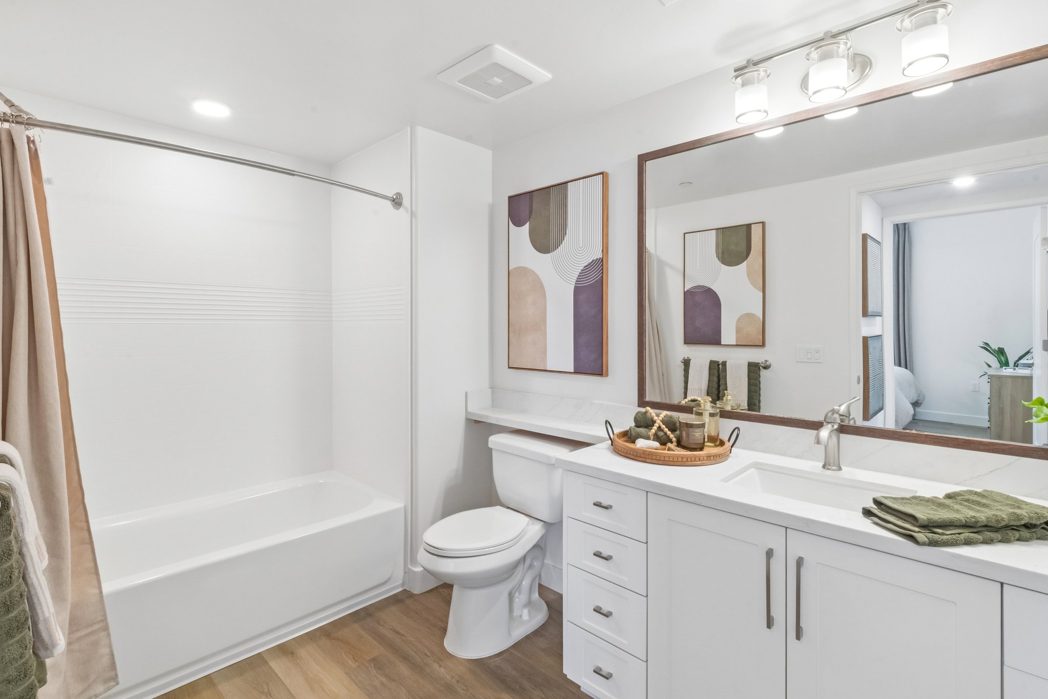 A modern bathroom featuring a bathtub with a shower, a white vanity with a sink, and a mirror above the sink. The decor includes two framed abstract art pieces on the wall and a basket with toiletries on the countertop. Soft lighting and neutral colors create a clean, inviting atmosphere.