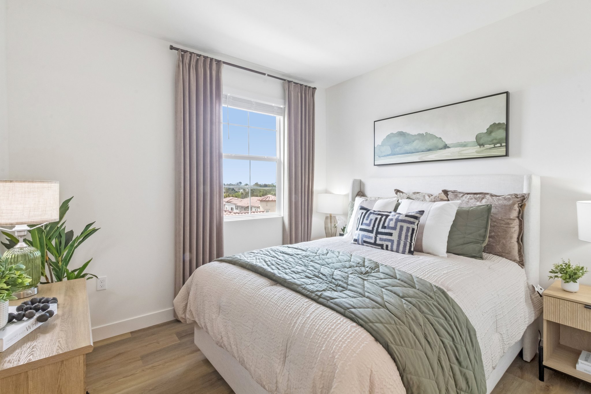Cozy bedroom featuring a comfortable bed with decorative pillows, soft bedding, and a quilt. There are two bedside lamps, a small plant, and a wooden nightstand. A large window allows natural light to enter, and a landscape painting is hanging on the wall. Light-colored walls enhance the airy feel of the space.