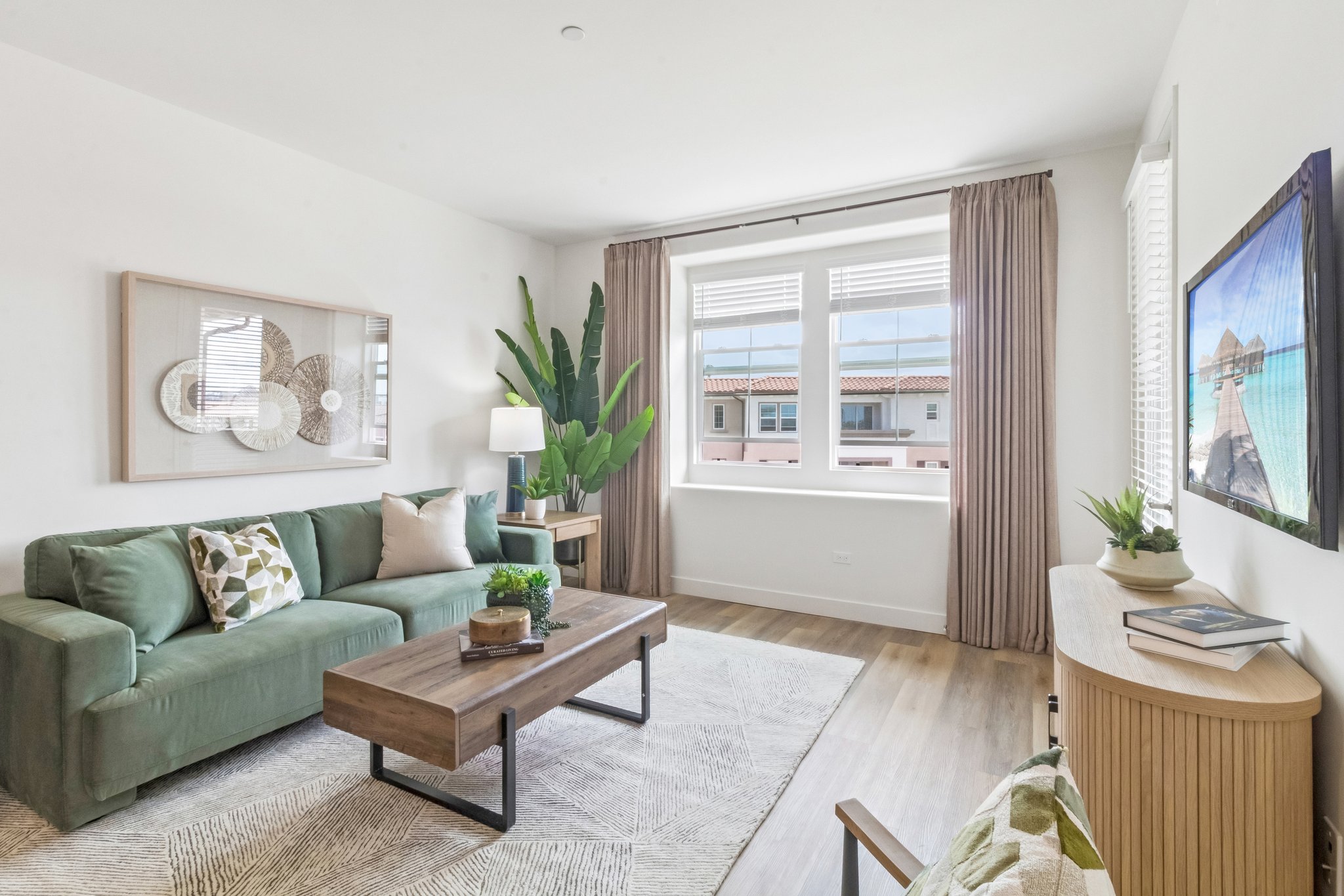 A stylish living room featuring a green couch with decorative pillows, a wooden coffee table, and a large window with curtains. A potted plant adds a touch of greenery, and there's a light wood sideboard and a framed picture on the wall. The overall decor is modern and inviting.