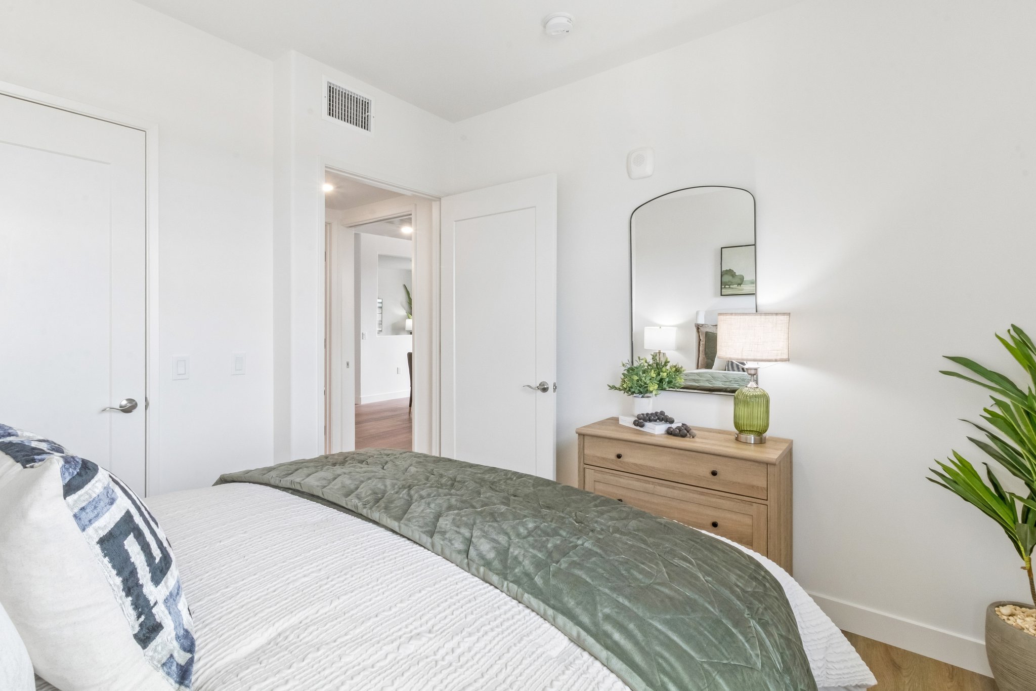 A modern bedroom featuring a neatly made bed with a green quilt, a wooden dresser, and a mirror. Soft lighting from a lamp, a decorative plant, and a light, neutral color palette create a calm and inviting atmosphere. The room is spacious with stylish decor and contemporary furnishings.