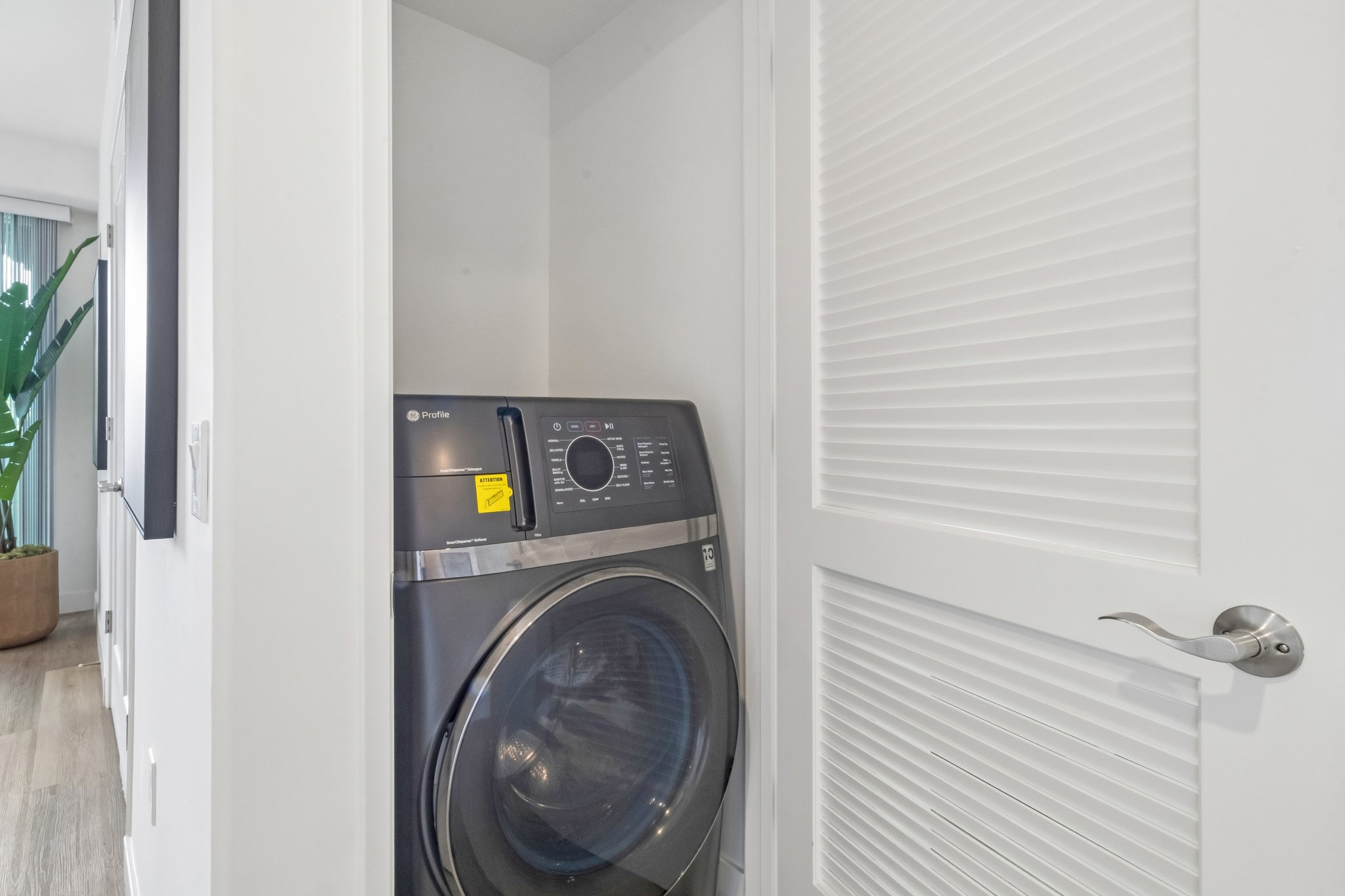 A modern washer and dryer tucked into a compact space, with a sleek design and control panel visible. The laundry area is framed by a white wall and a door, suggesting a tidy, organized interior. A potted plant can be seen in the background, adding a touch of greenery to the setting.