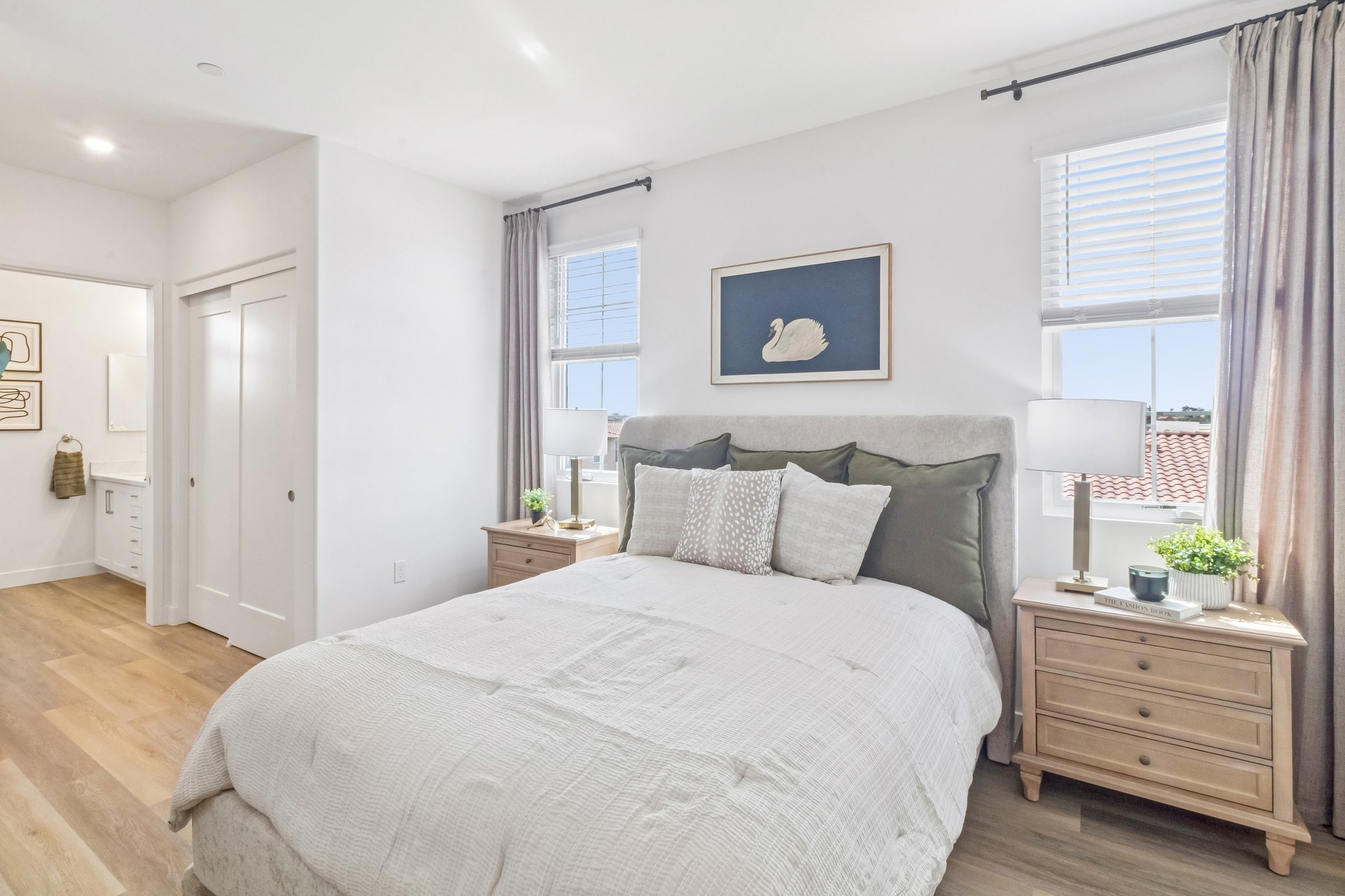 A modern bedroom featuring a queen-sized bed with a light gray upholstered headboard and decorative pillows in neutral tones. Two wooden nightstands with plants are placed on either side, and large windows allow natural light to fill the space. In the background, there's a doorway leading to a bathroom.