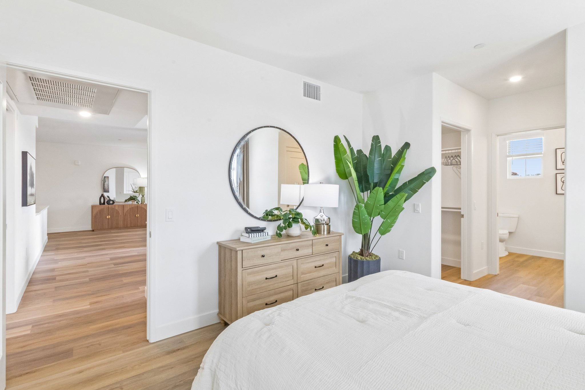 A bright and modern bedroom featuring a large bed with white bedding, a round mirror above a wooden dresser, and a potted plant. The room has light wood flooring and white walls, with an open doorway leading to a hallway and an adjacent bathroom visible in the background.