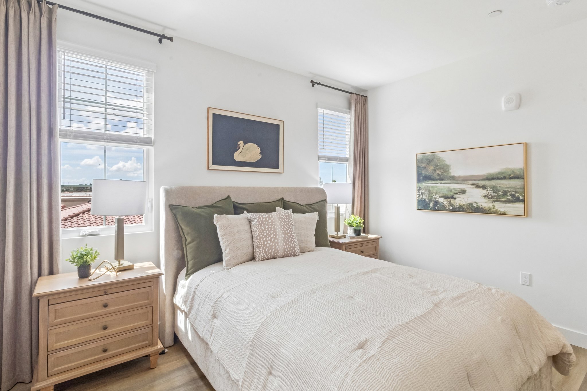 A cozy bedroom featuring a queen-sized bed with decorative pillows, light-colored bedding, and elegant headboard. Two bedside tables with potted plants and lamps are on either side. The room has large windows with sheer curtains, letting in natural light, and framed artwork depicting a serene landscape is on the wall.
