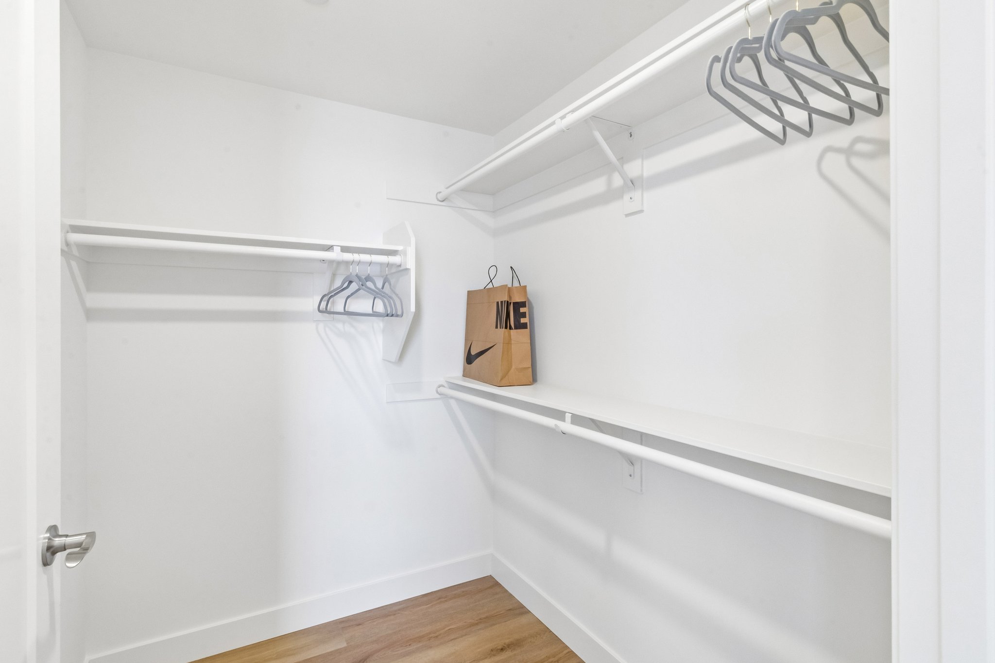 Empty closet space featuring a white wall and wooden floor. There are two clothing rods with several hangers on one side and a single paper shopping bag from Nike on the floor. The overall design is minimalist with ample storage potential.