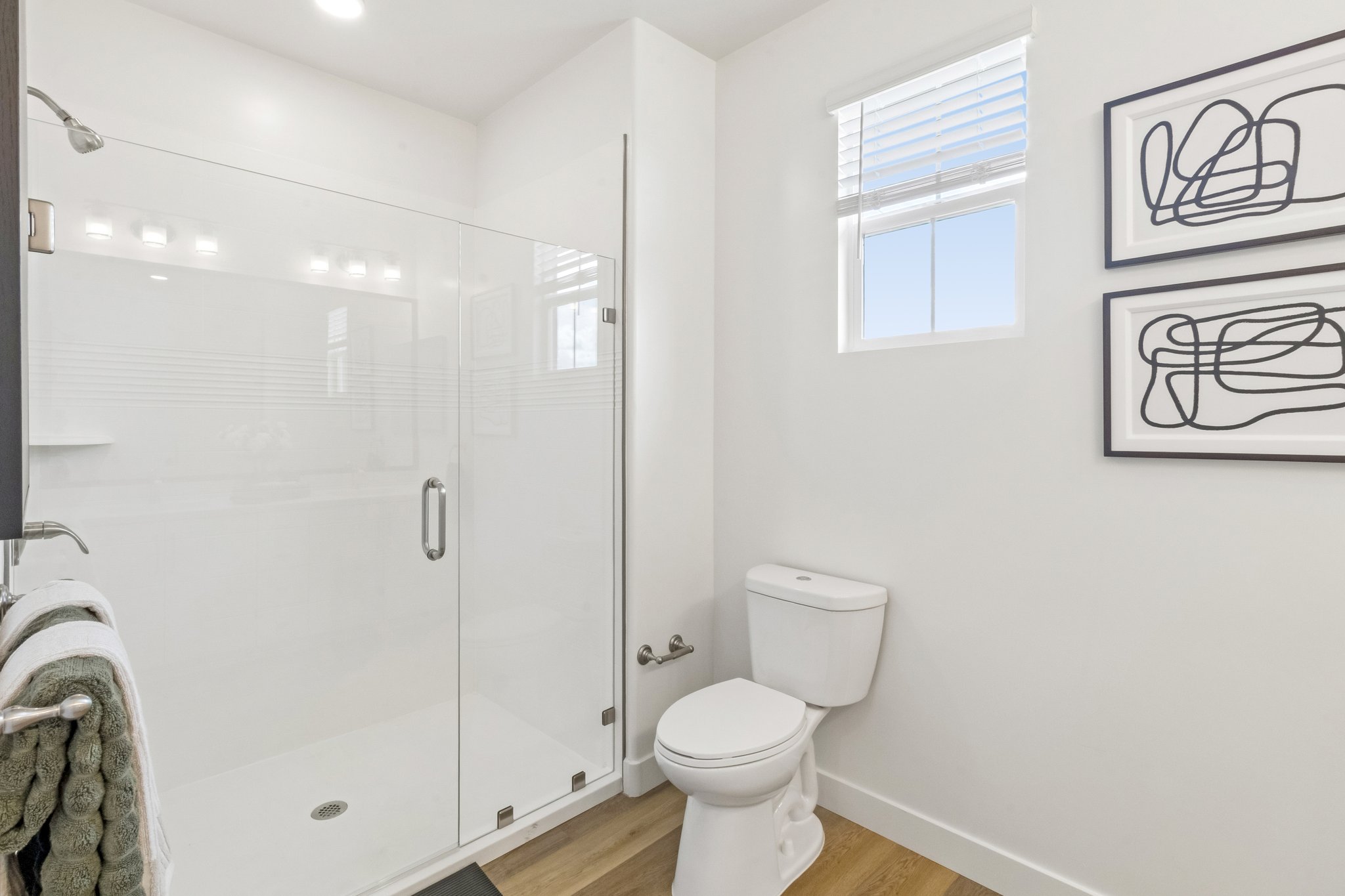 A modern bathroom featuring a glass-enclosed shower with a sleek design, a white toilet, and light wood flooring. Two abstract framed artworks hang on the wall, with a window allowing natural light to enter the space. Towel hanging on a rack adds functionality and comfort.