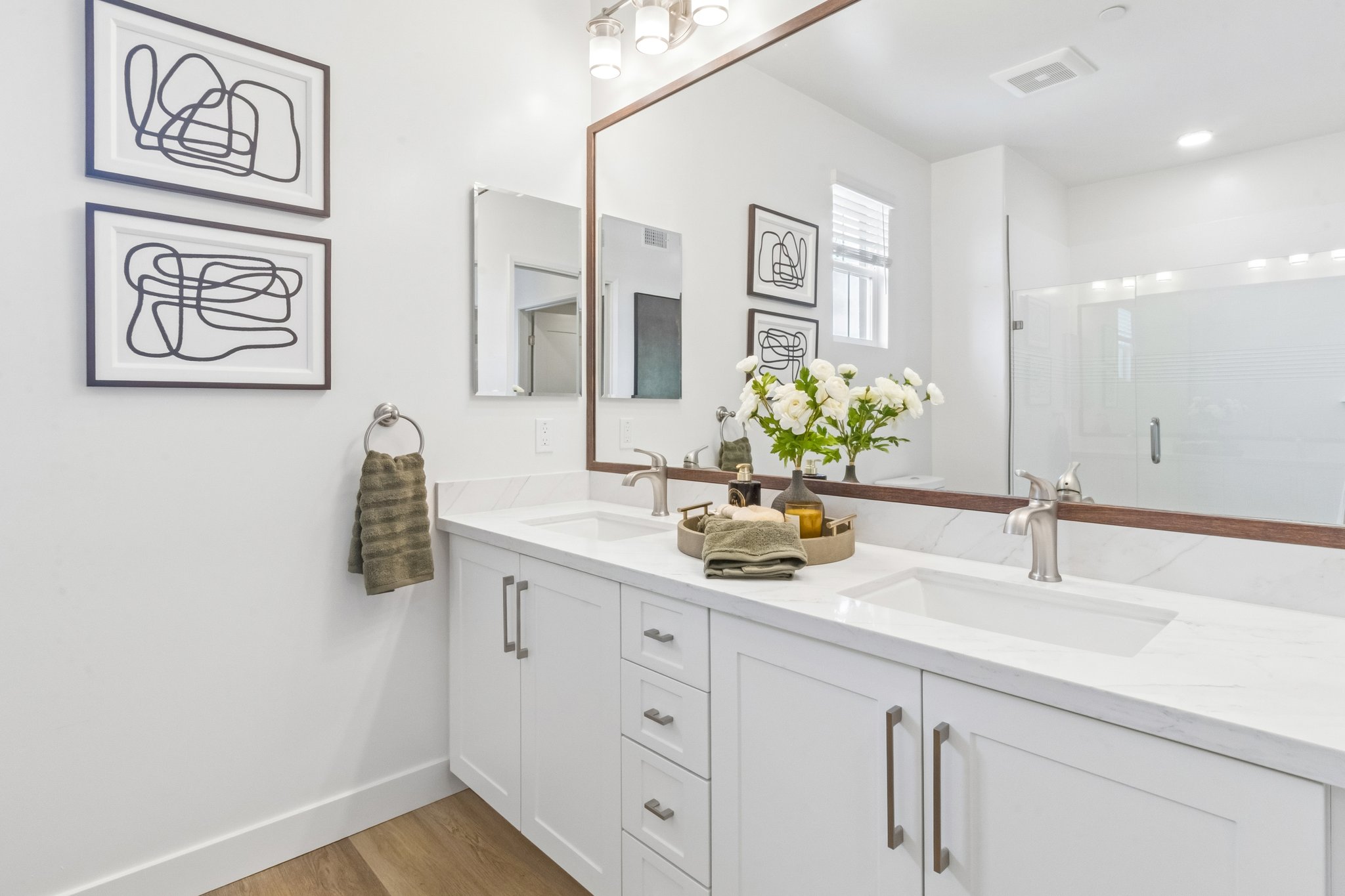 A modern bathroom with a double vanity featuring a marble countertop. Above the sinks are two abstract framed artworks. A round mirror reflects the space, and a bouquet of white flowers is in a wooden tray. Towel and toiletries are neatly arranged, and there is a large glass shower in the background.