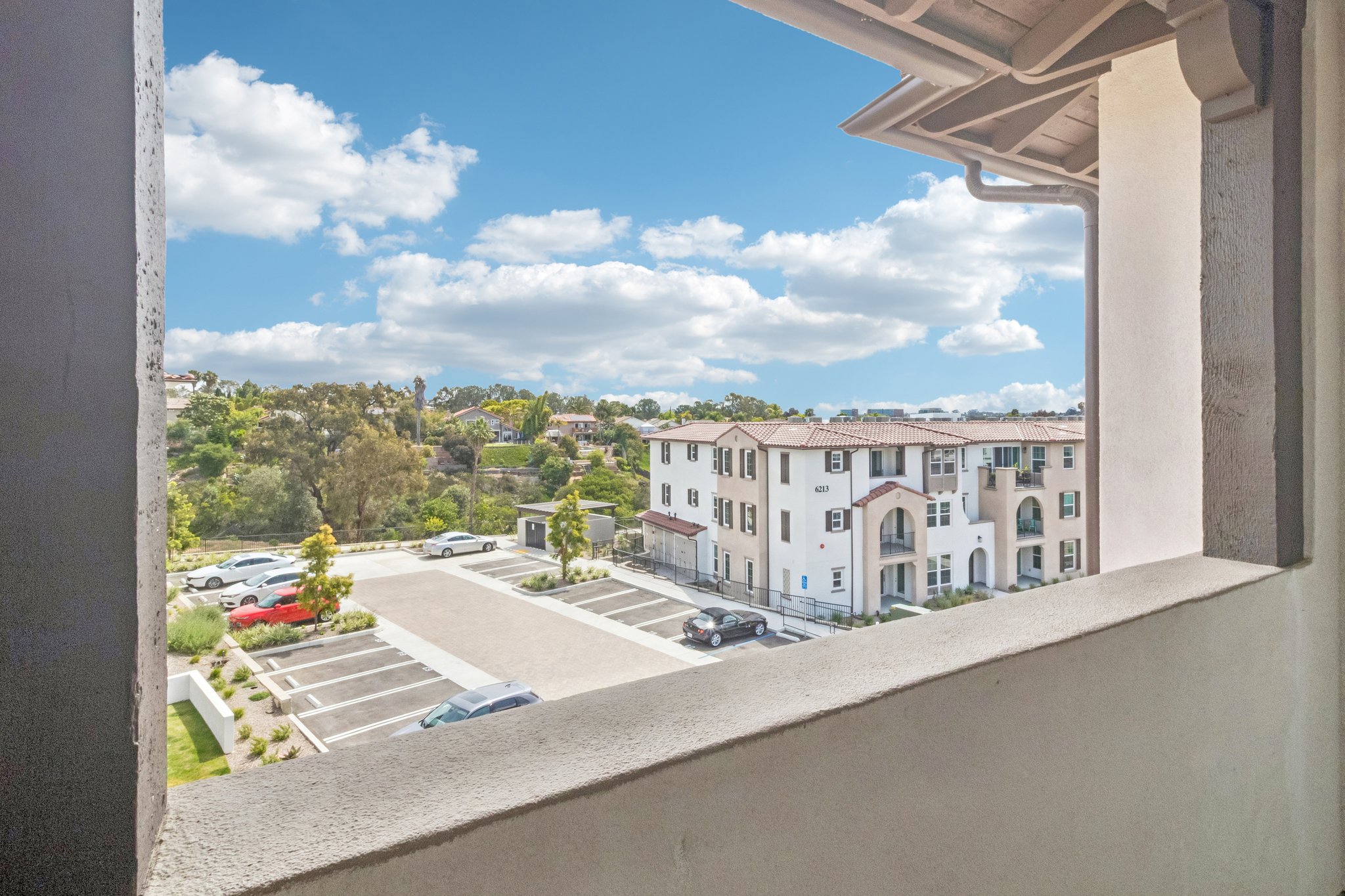 View from a window showing a parking lot and residential buildings. Lush green trees and a clear blue sky with fluffy white clouds can be seen in the background, creating a serene suburban atmosphere.