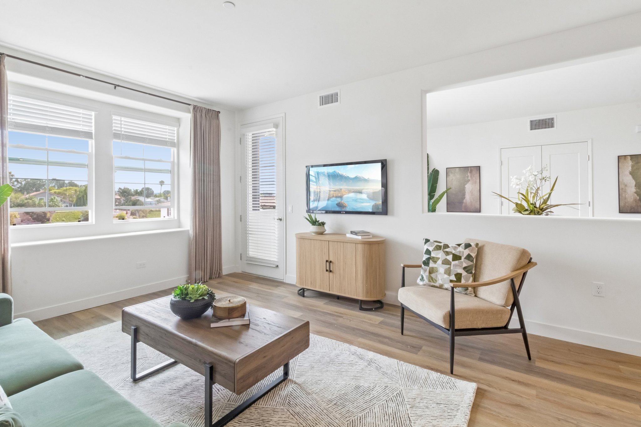 A modern living room featuring a light-colored sofa, a wooden coffee table with a plant, and an armchair. There's a television on a cabinet against the wall, large windows letting in natural light, and soft curtains. Decorative elements include a rug and a small indoor plant.