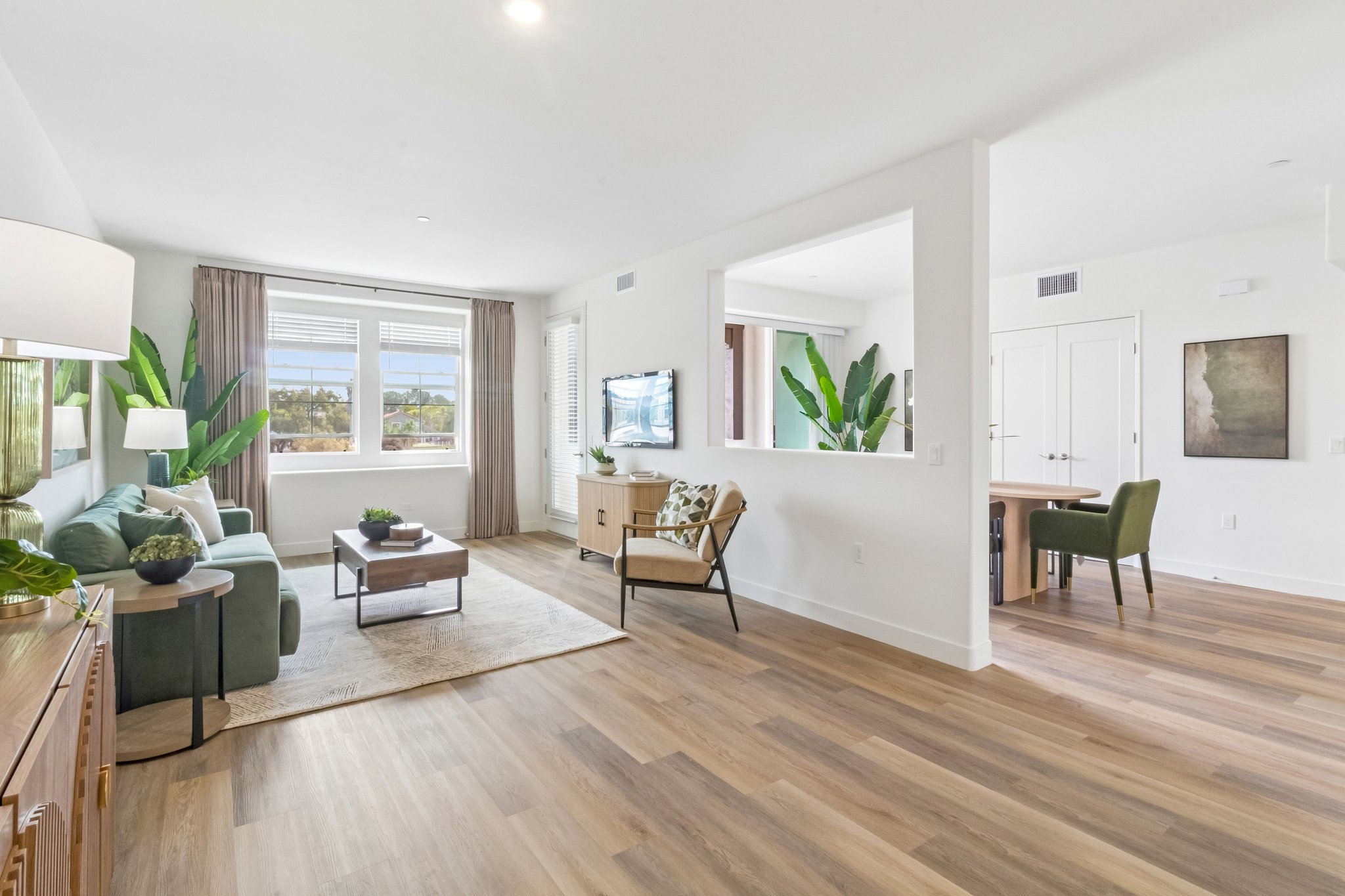 A well-furnished living room featuring a green couch, a coffee table, and a large window with natural light. In the background, a dining area with a table and chairs, and decorative indoor plants are visible. The flooring is light wood, creating a warm, inviting atmosphere.