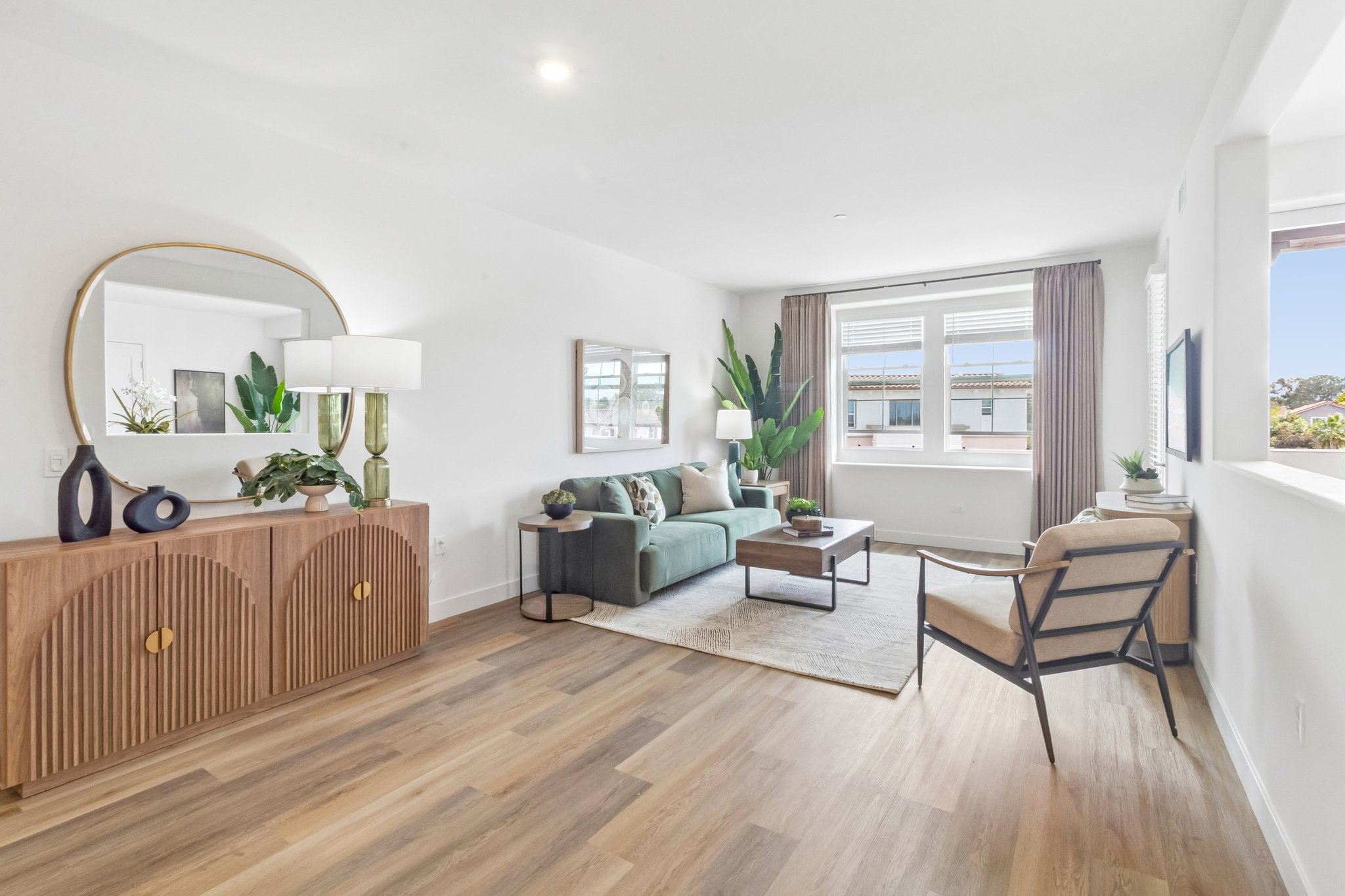 A modern living room featuring a green sofa, a wooden coffee table, and a stylish armchair. Natural light floods the space through large windows, with potted plants adding a touch of greenery. A large mirror and minimalist decor enhance the contemporary feel of the room.