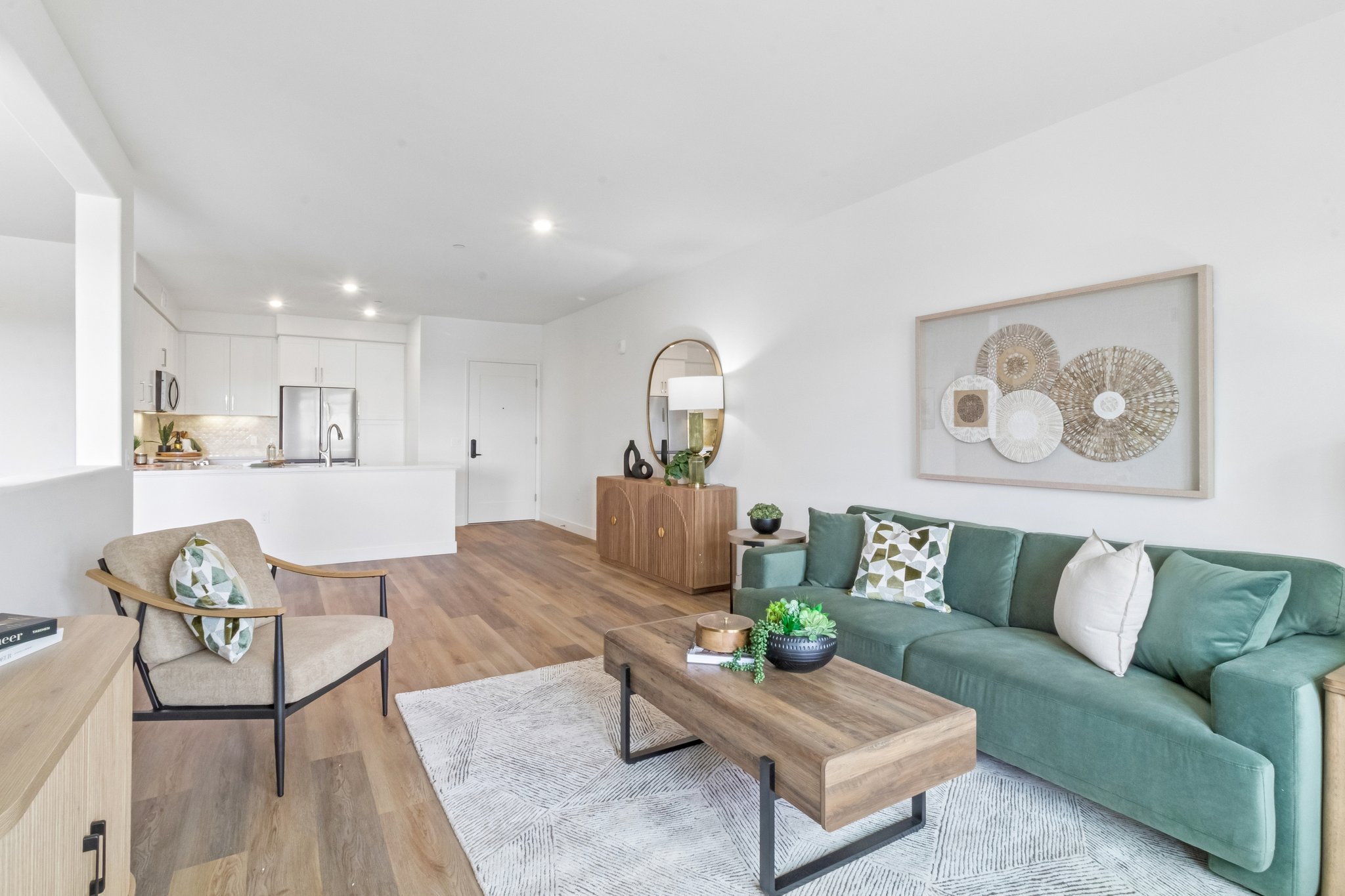 A modern living room featuring a green sofa with decorative cushions, a wooden coffee table, and a patterned rug. A beige armchair is positioned nearby. In the background, there is a kitchen area with sleek cabinetry and a round mirror on the wall, enhancing the contemporary aesthetic.