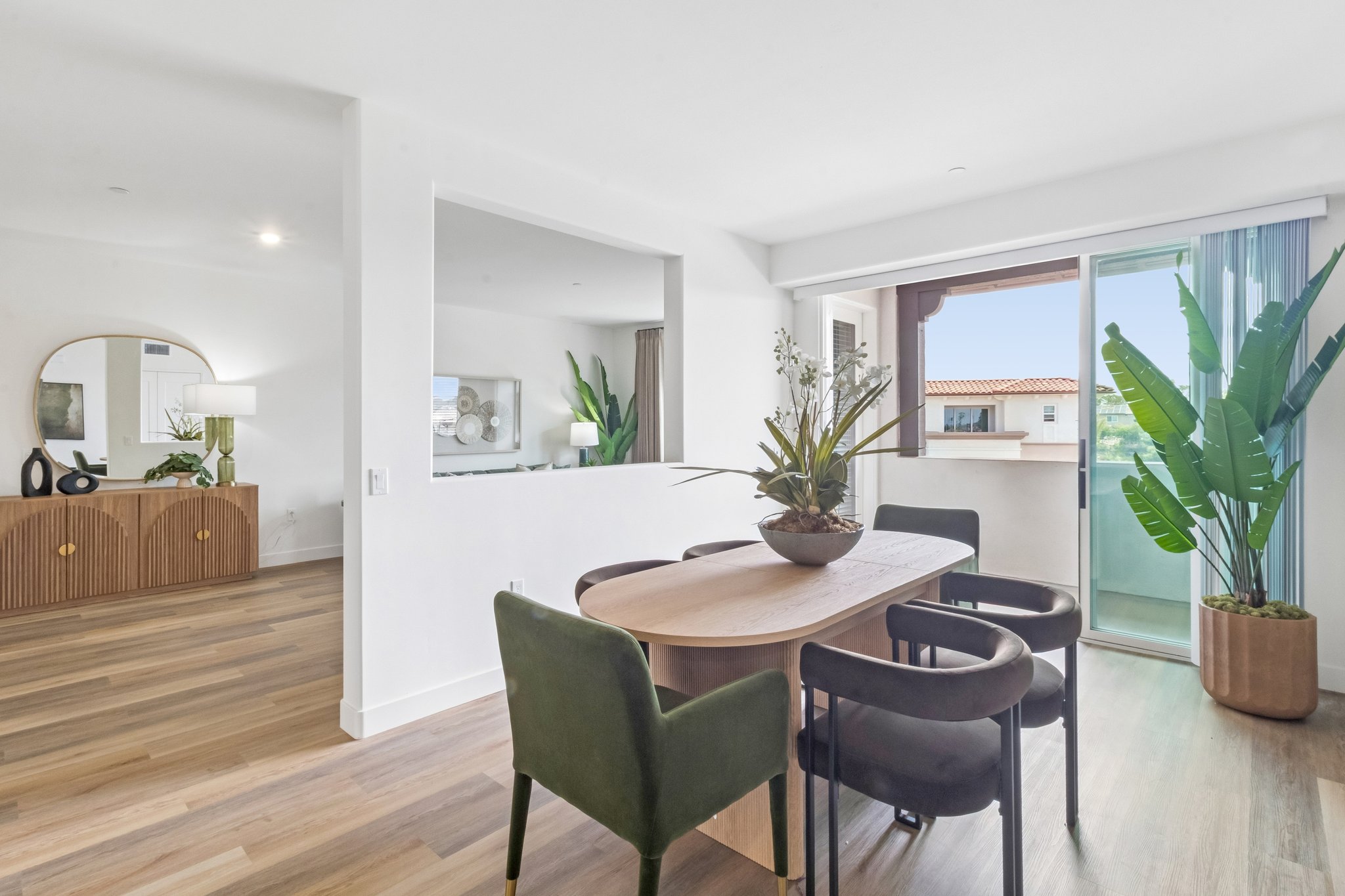 A modern dining area featuring a round wooden table surrounded by green chairs. A large planter with a leafy plant sits at the center of the table. In the background, there's a spacious living area with a stylish sideboard and a wall of windows showcasing a view of the outdoors.