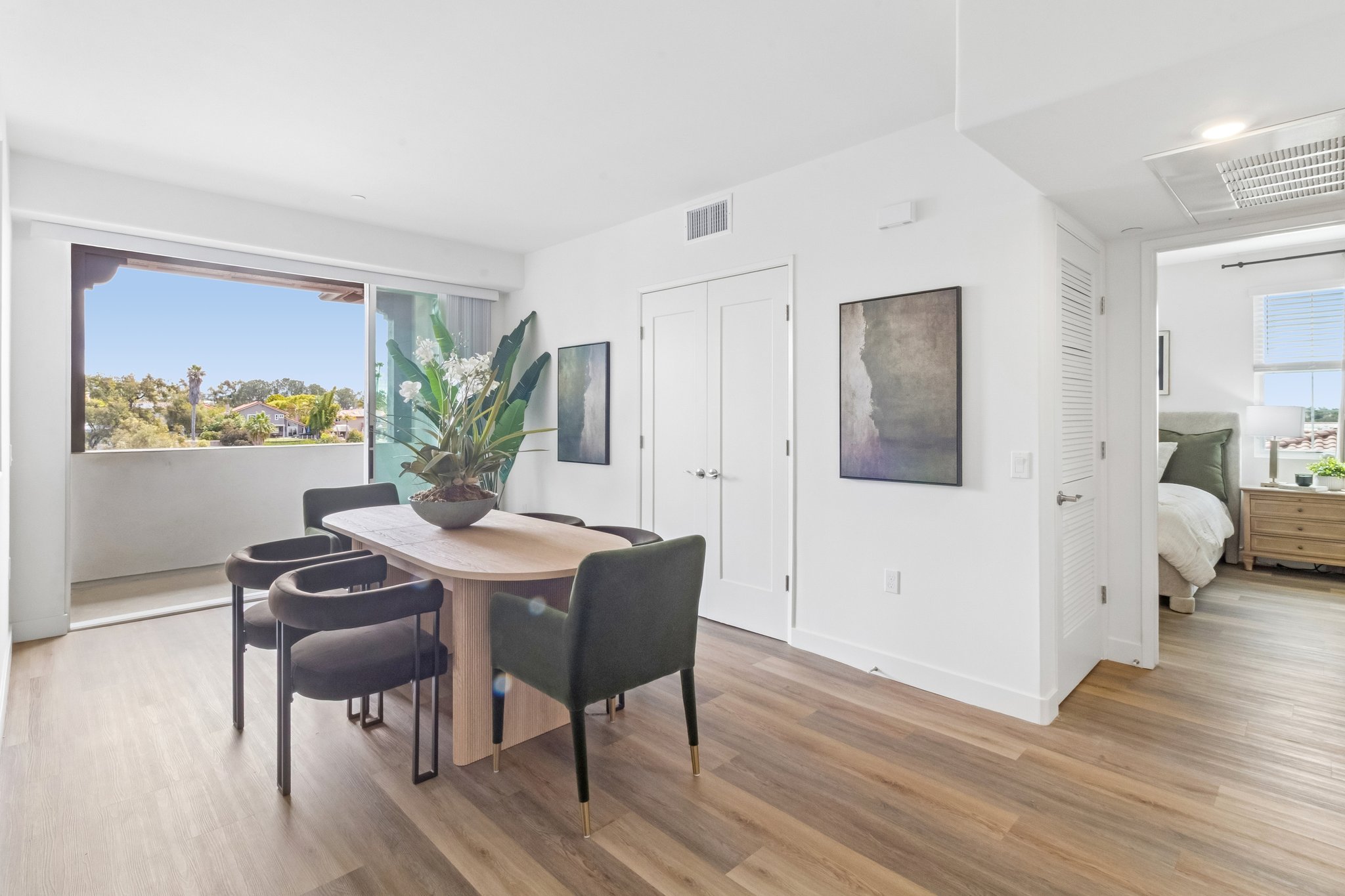 A modern, brightly lit interior featuring a dining area with a wooden table and four black chairs. A large plant in a vase is placed on the table. The space includes a doorway leading to a balcony with a view, and two framed artworks on the walls. The adjacent room appears to be a bedroom.