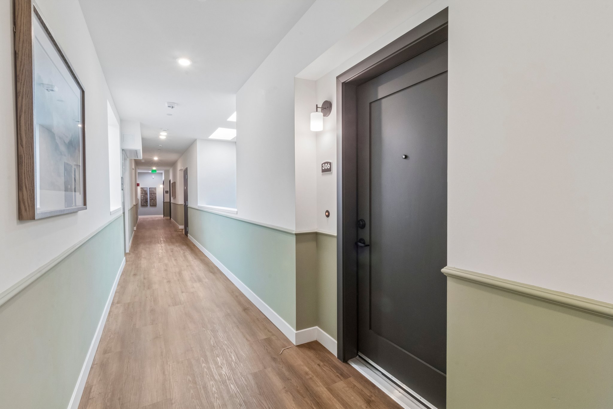 A modern hallway featuring light wood flooring, pale green and white walls, and a dark door at the end. The space is well-lit with natural light and overhead lights, and there are framed artworks on the walls, enhancing the contemporary decor.