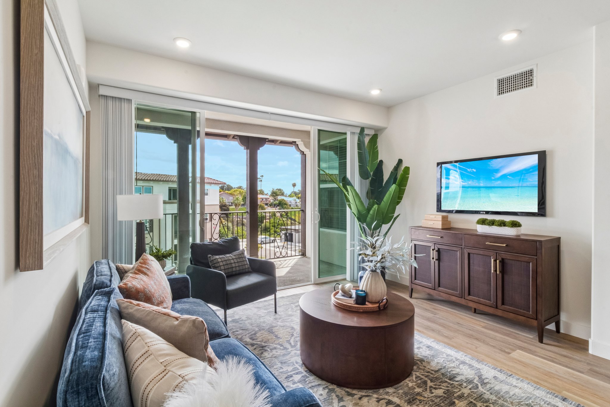 A modern living room featuring a blue sofa, a round coffee table, and a wooden TV cabinet. There are large sliding glass doors opening to a balcony with a view, and a potted plant adds greenery to the space. The room is brightly lit with natural light and has a cozy, inviting atmosphere.