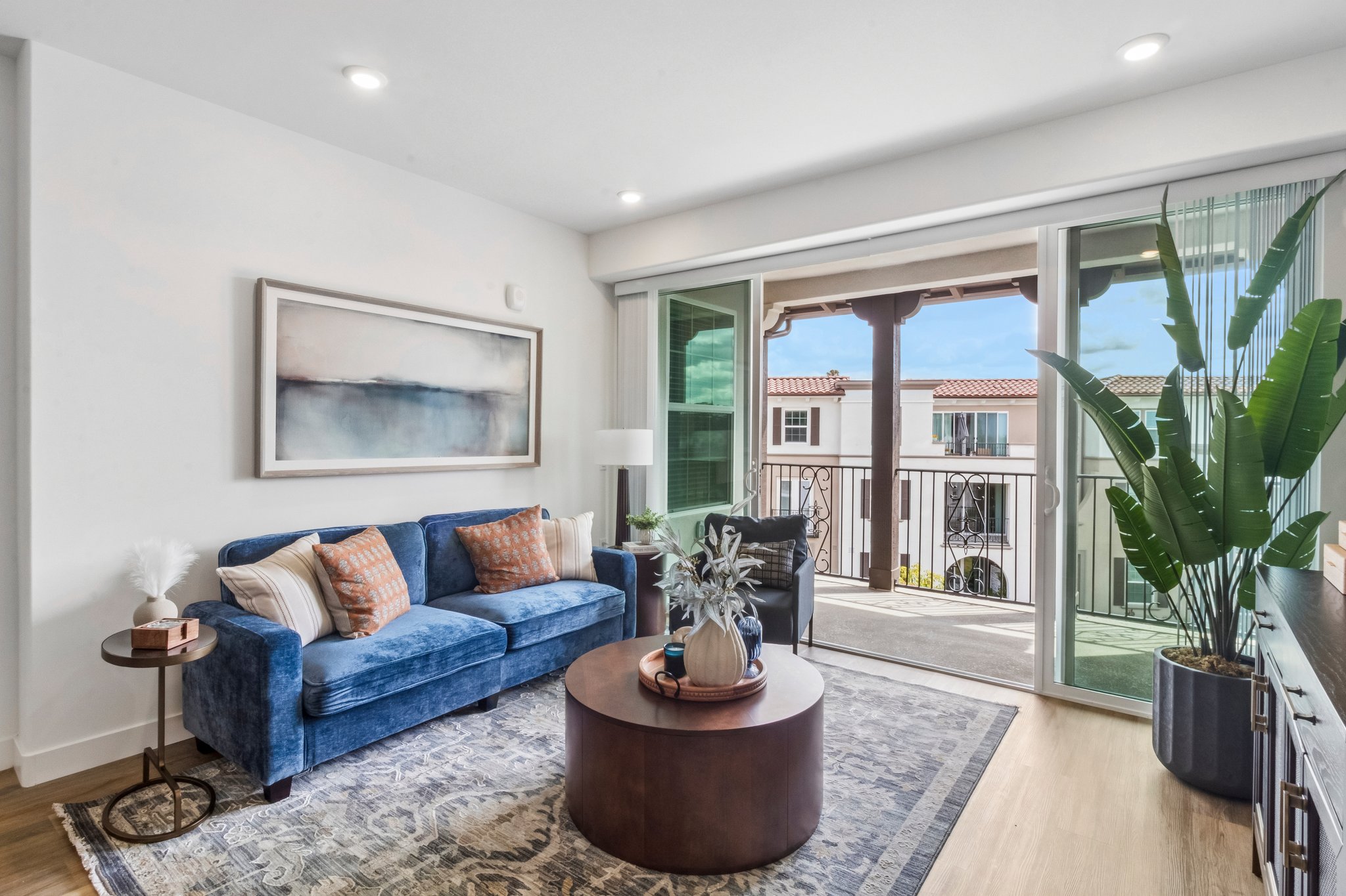 A modern living room featuring a blue sofa with decorative pillows, a round coffee table, and a large potted plant. Glass sliding doors open to a balcony with a view of neighboring buildings. The room has neutral walls and elegant decor, creating a cozy and inviting atmosphere.
