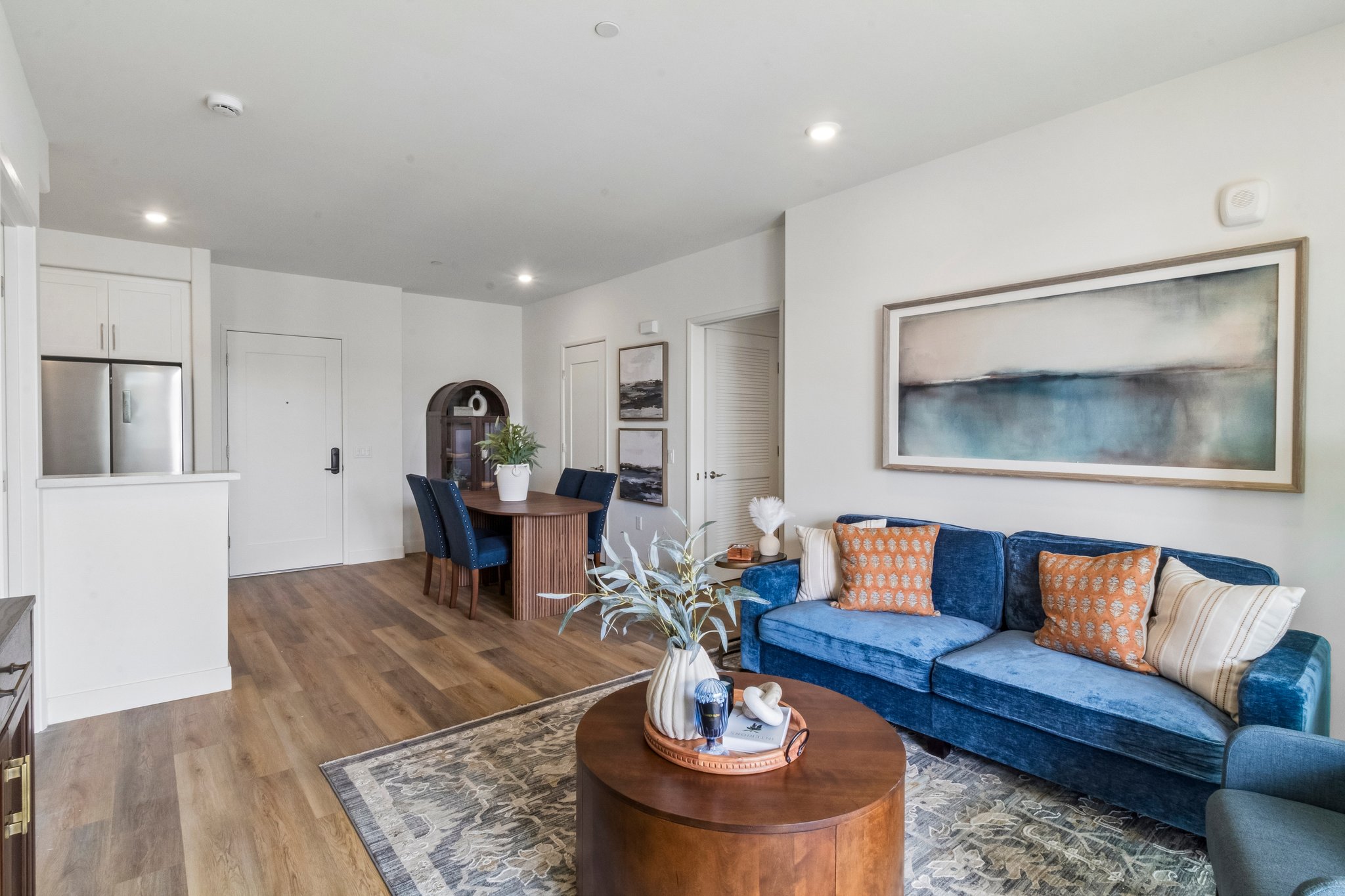 A modern living room featuring a stylish blue sofa with patterned cushions, a round wooden coffee table, and a decorative plant. The space includes a dining area with a small table and chairs, an open kitchen with stainless steel appliances, and large artwork on the wall, all set in a light, airy atmosphere.