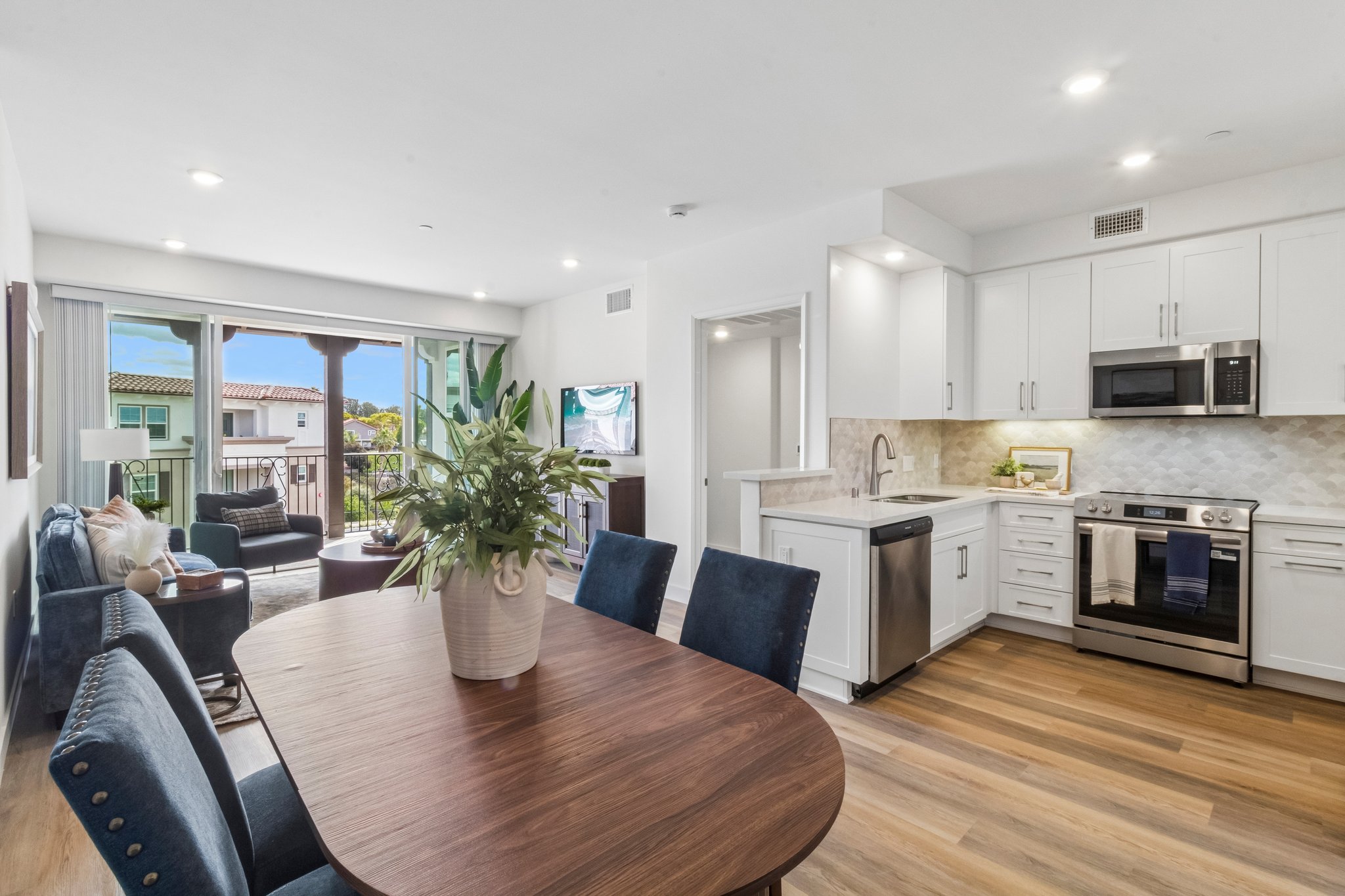A modern kitchen and dining area featuring a wooden table with blue upholstered chairs. The kitchen has white cabinets, stainless steel appliances, and a light stone backsplash. Large windows allow natural light in, showcasing a view of greenery outside.
