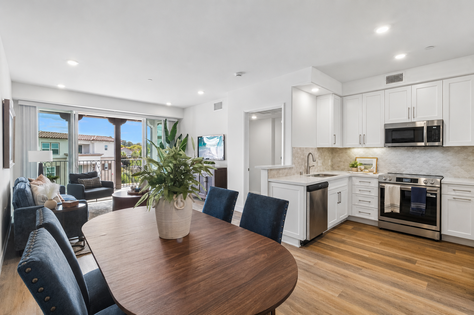 Modern kitchen and dining area featuring white cabinetry, stainless steel appliances, and a wooden dining table with blue chairs. A large window provides natural light, while a balcony can be seen in the background. The decor is contemporary and inviting.