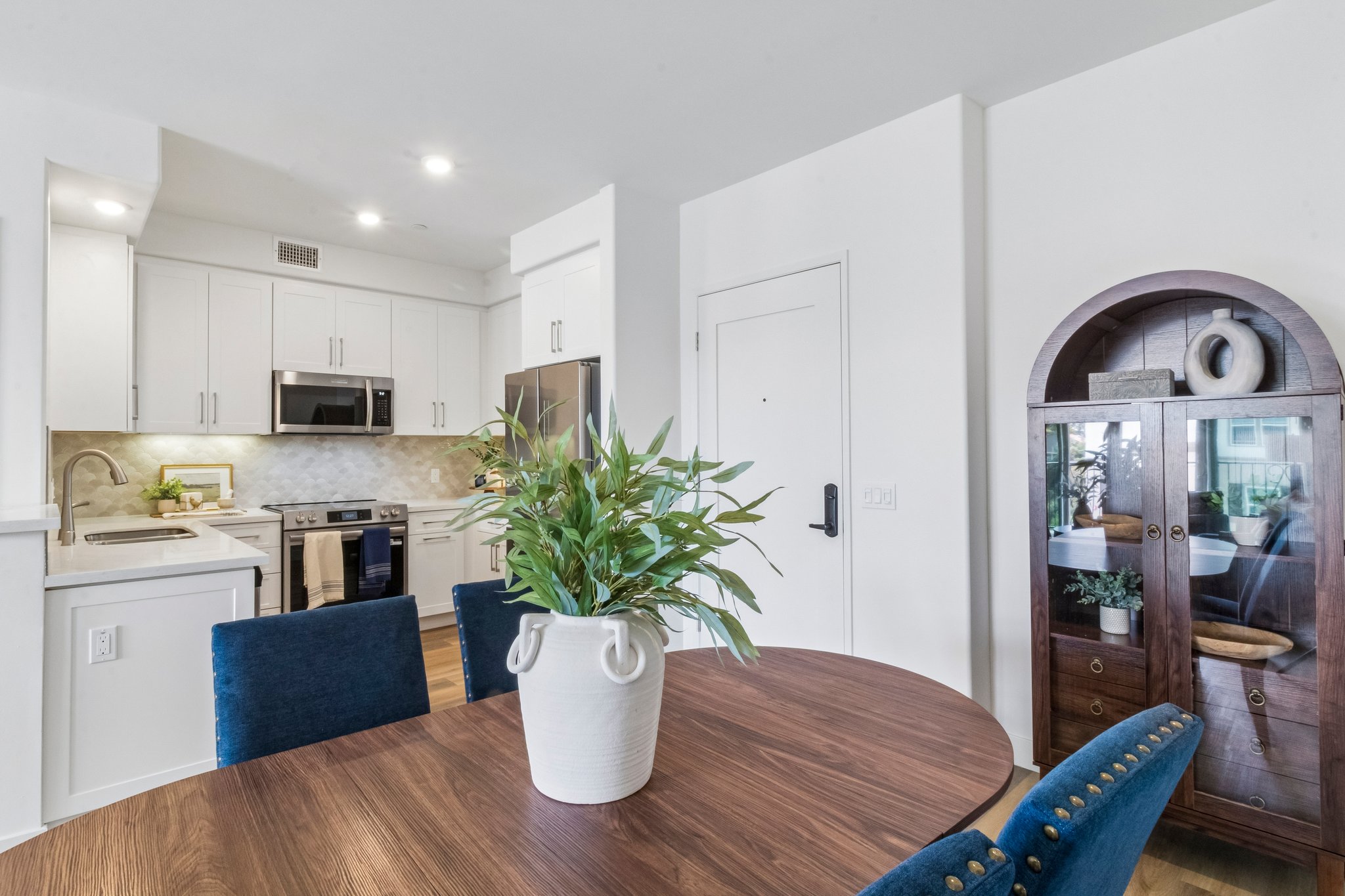 Modern kitchen and dining area featuring a round wooden table with blue upholstered chairs. A large potted plant sits at the center of the table. The kitchen showcases white cabinetry, stainless steel appliances, and a tiled backsplash. A decorative wooden cabinet is visible in the corner, adding warmth to the space.