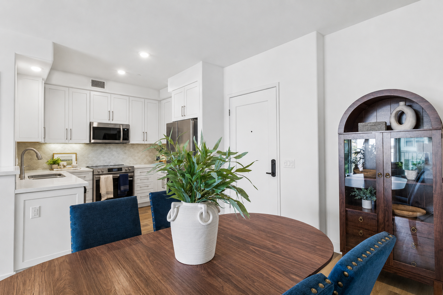 A modern kitchen and dining area featuring a round wooden table with blue upholstered chairs. A large white planter with green plants sits on the table. The kitchen has white cabinets, stainless steel appliances, and a light backsplash. A decorative wooden cabinet with glass doors is visible in the background.