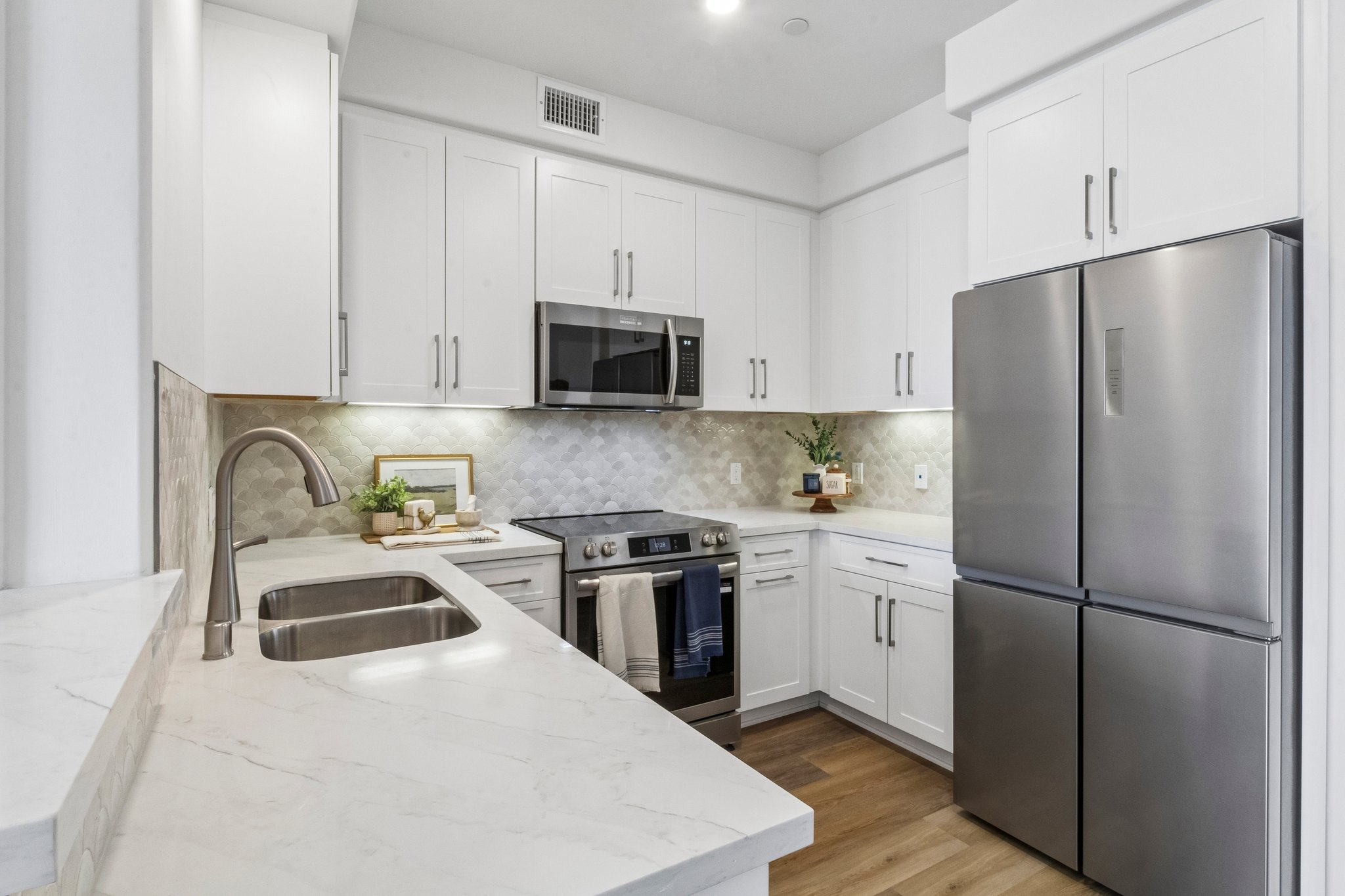Modern kitchen featuring white cabinetry, stainless steel appliances including a microwave and refrigerator, a silver sink, and a marble countertop. The backsplash has a textured design, and there's a small plant on the countertop, creating a clean and inviting space.