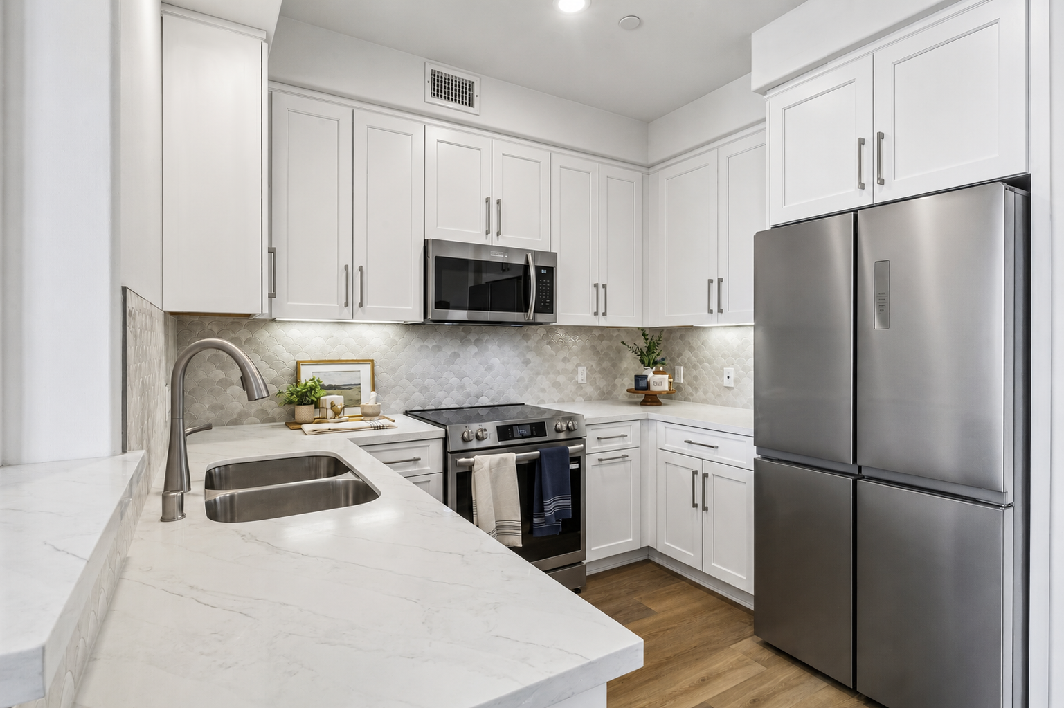 Modern kitchen featuring white cabinetry, a stainless steel refrigerator, and a gas stove. The countertop is made of marble, and the backsplash is tiled in a light gray pattern. A sink is visible, and decorative plants and kitchen items are arranged on the countertop. Natural light illuminates the space.