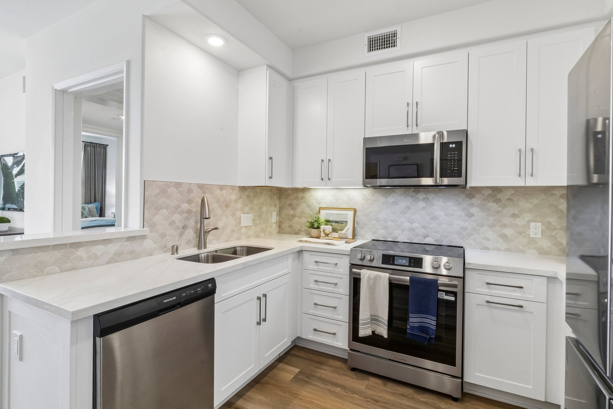 Modern kitchen featuring white cabinets, a stainless steel refrigerator, built-in microwave, and an oven range. The countertop is made of light-colored stone, and there is a sink with a geometric tile backsplash. Soft lighting enhances the contemporary design, creating a bright and inviting atmosphere.