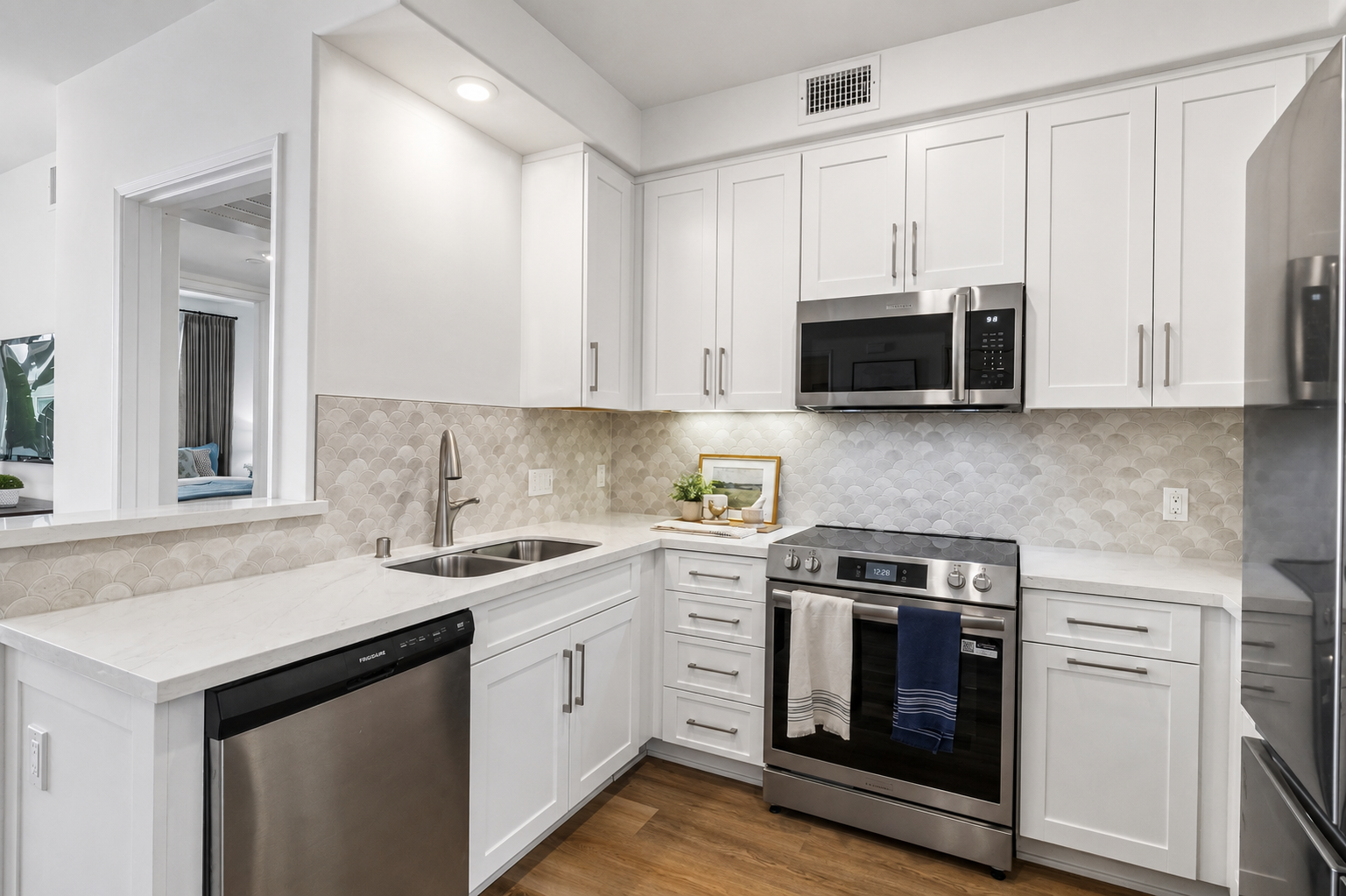 Modern kitchen featuring white cabinetry, a stainless steel refrigerator, microwave, and a black oven with a stovetop. The countertop is white with a subtle veining pattern, and there is a stainless steel sink. The backsplash has a textured tile design, and a small plant and framed picture add decorative touches.