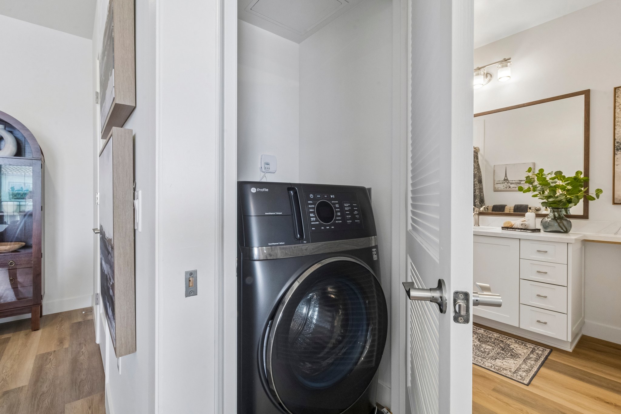 A modern laundry area featuring a front-loading washing machine situated between a bathroom and hallway. The machine is black with a digital control panel, and the space is light and airy with wooden floors and subtle decor elements like a mirror and plant.