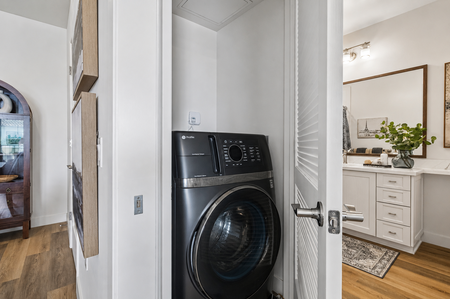 A modern laundry area features a black front-loading washing machine positioned in a compact space. The doorway leads to a stylish bathroom with contemporary fixtures, a large mirror, and decorative elements like a plant and artwork on the walls, highlighting a blend of functionality and aesthetics.