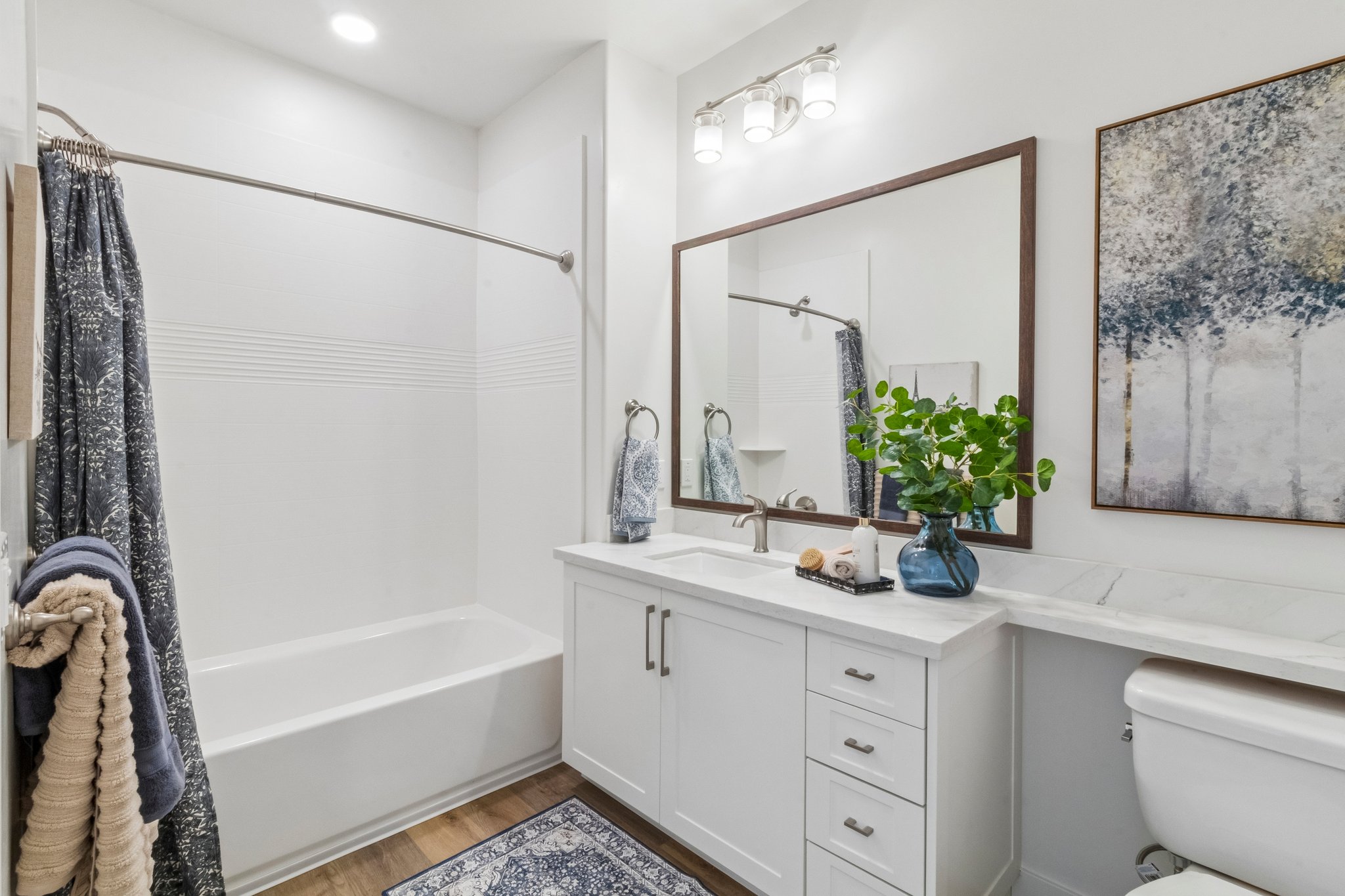 A modern bathroom featuring a bathtub with a curved shower curtain rod, a double sink vanity with a stylish mirror, and natural light from ceiling fixtures. Decorative elements include a plant on the counter, towels hanging, and a piece of art on the wall, enhancing the space's contemporary design.
