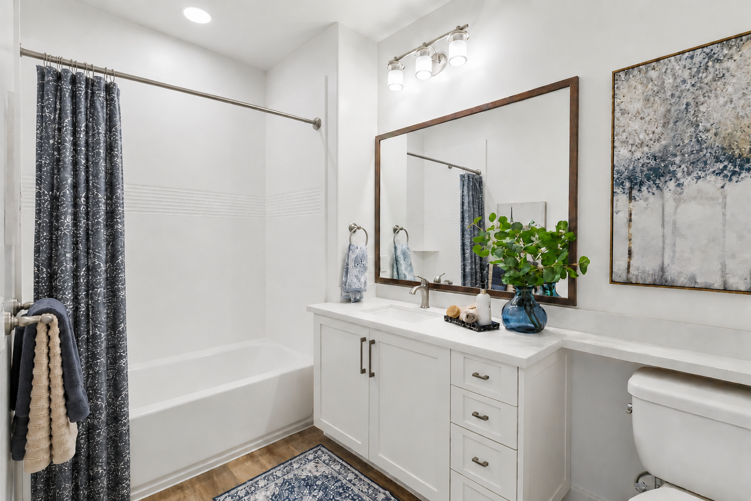 A modern bathroom featuring a white bathtub with a shower curtain, a double vanity with a large mirror, a decorative painting, and a small plant. The space is bright and features wooden flooring and stylish fixtures, creating a clean and inviting atmosphere.