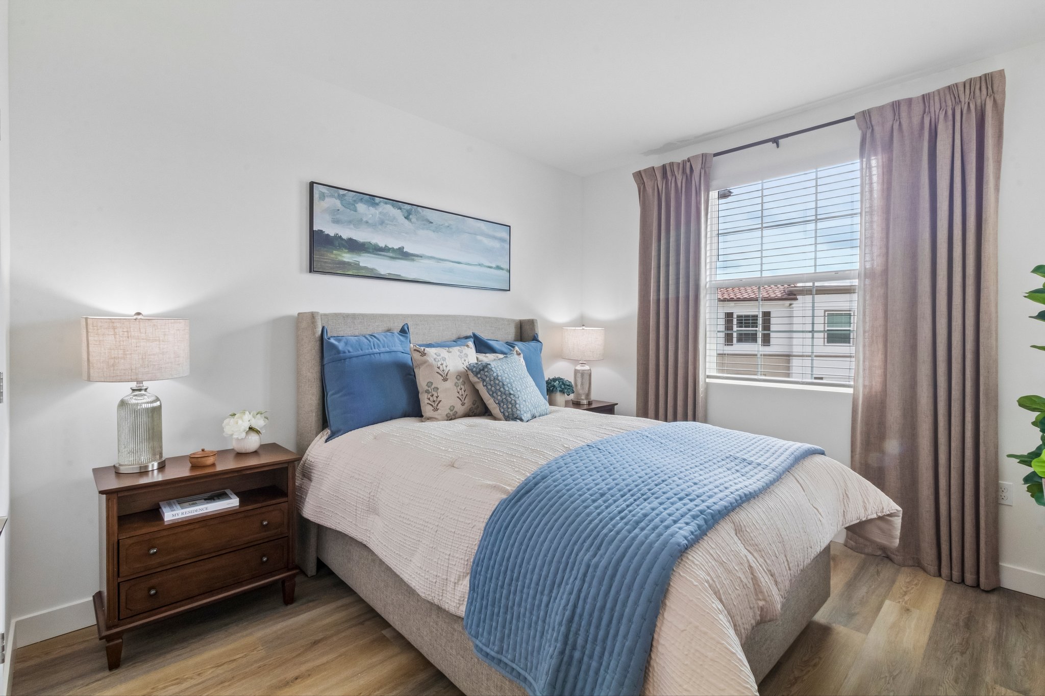 A cozy bedroom featuring a bed with blue and beige bedding, two lamps on bedside tables, and decorative pillows. A painting of a landscape hangs above the bed. Natural light streams in through a window covered with beige curtains, while a potted plant adds a touch of greenery to the space.
