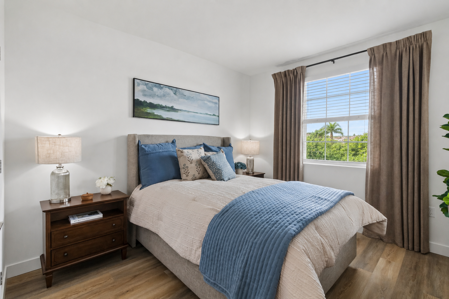 A cozy bedroom featuring a comfortable bed with blue and white bedding, decorative pillows, and a light gray headboard. A wooden nightstand holds a lamp and a small vase of flowers. The room is bright with a window showcasing greenery outside, and soft curtains add a touch of elegance.