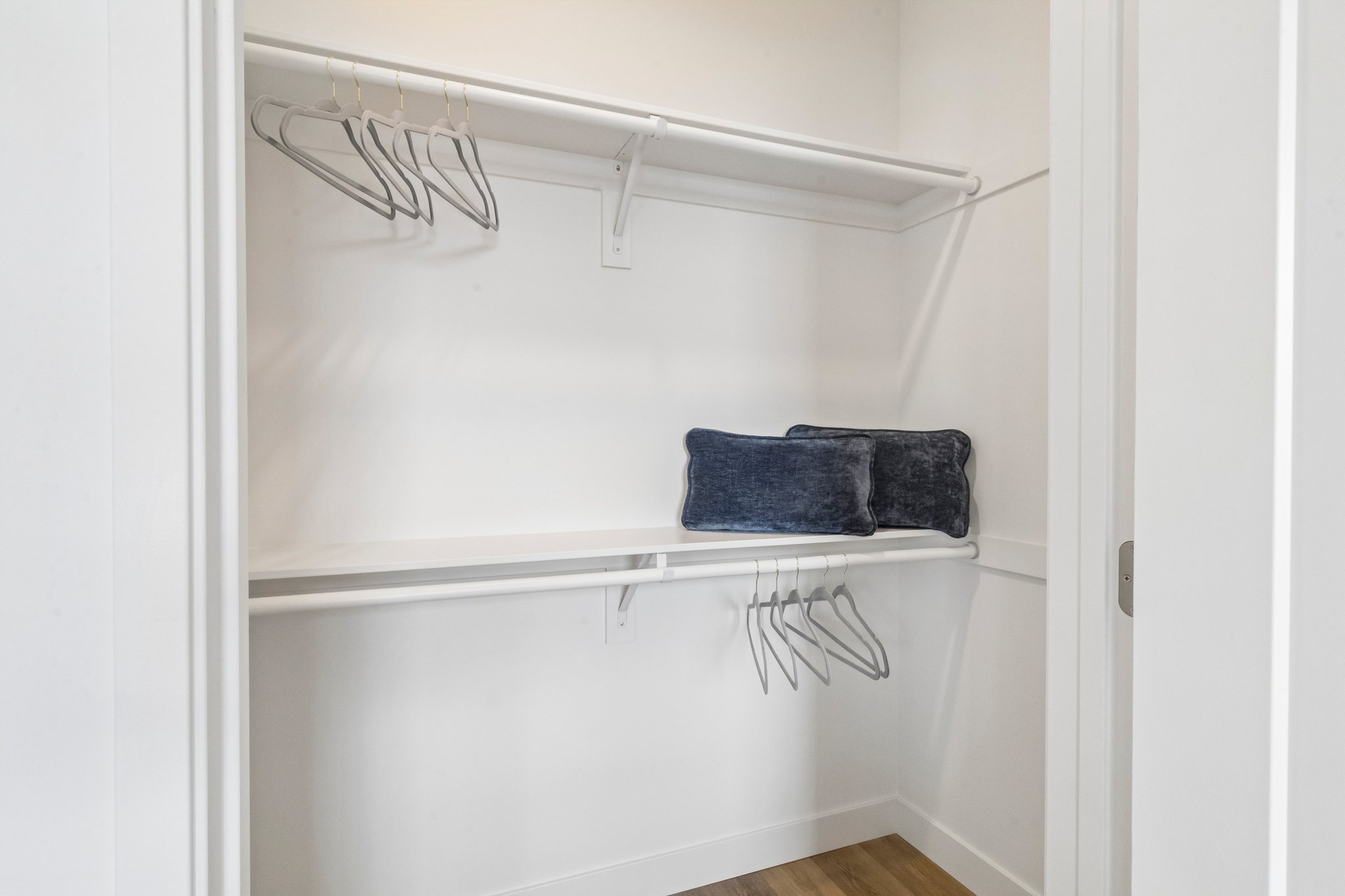 An empty closet with white walls and a wooden floor. It features a top shelf with hangers, a lower shelf, and two dark blue pillows. The closet is well-lit and organized, showcasing ample storage space.