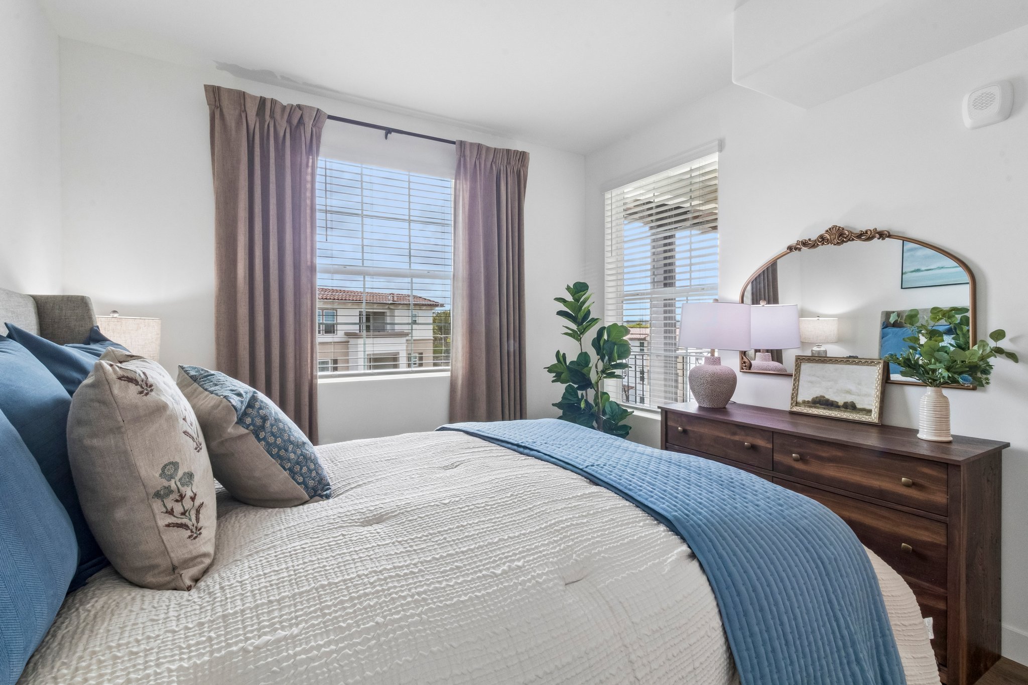 A cozy bedroom featuring a neatly made bed with blue and cream linens. There are decorative pillows on the bed, a wooden nightstand with a lamp and a framed photo, and potted plants. Natural light enters through a window with sheer curtains, showcasing an outside view.