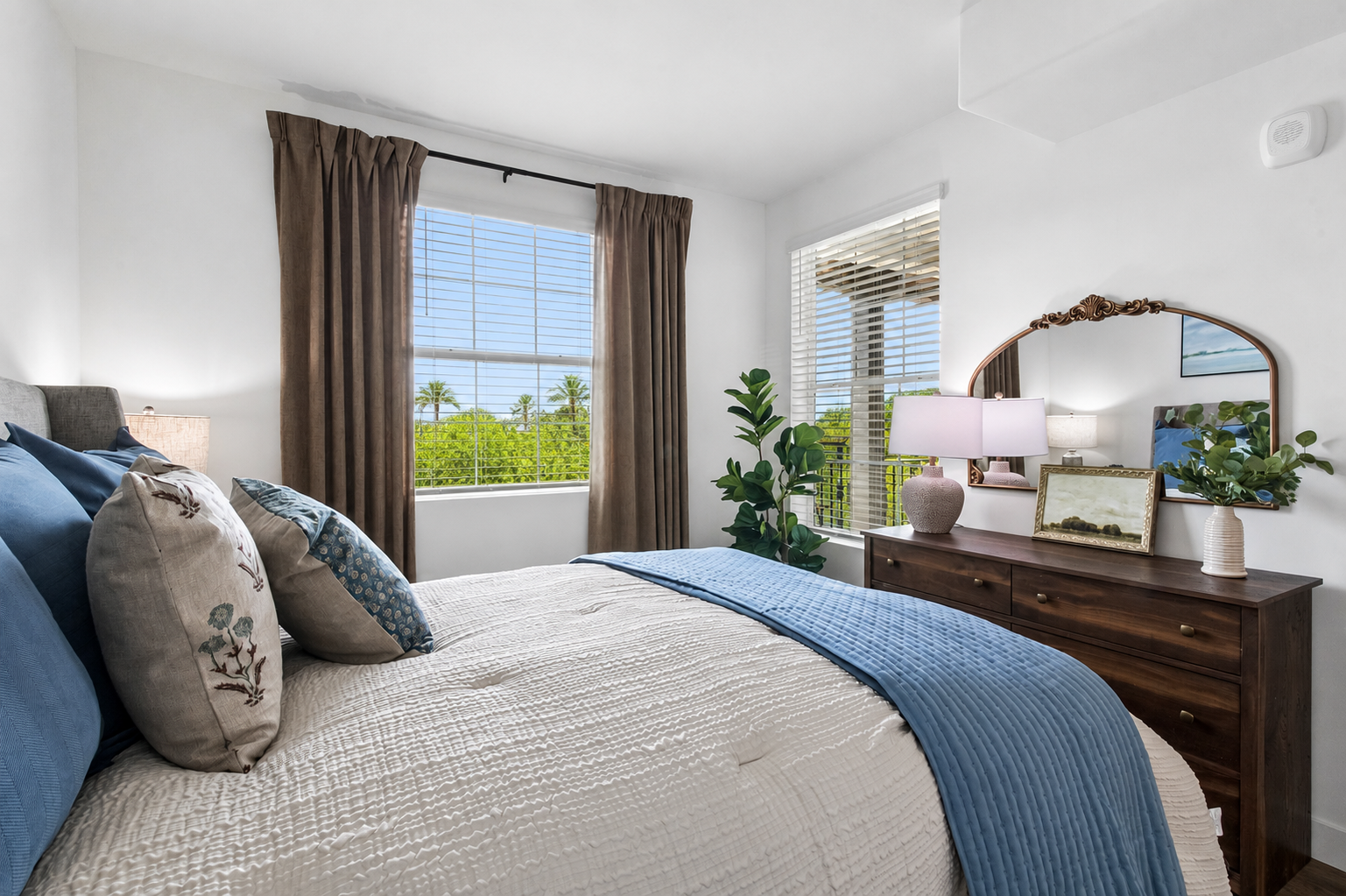 A cozy bedroom featuring a neatly made bed with blue and beige accents. There are curtains framing a window with a view of greenery, a bedside table with a lamp, and a wooden dresser with a decorative mirror and plants, creating a calm and inviting atmosphere.